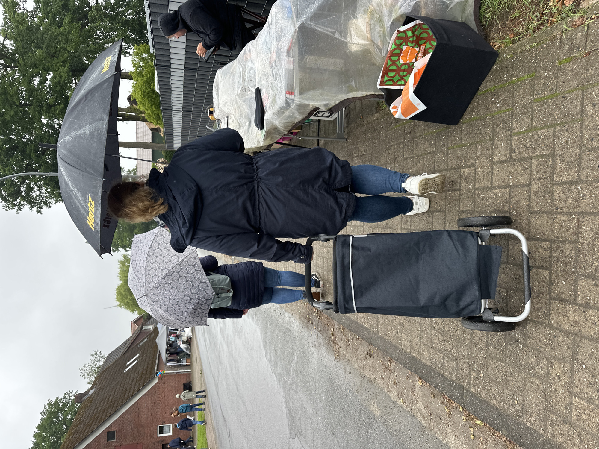 The image depicts an outdoor scene on a wet, overcast day in Deinste, Germany, likely a street market or yard sale. In the immediate foreground, a person with their back to the camera, wearing a dark blue hooded jacket and blue jeans, pushes a black fabric shopping trolley with two wheels on a grey brick sidewalk. To their left, a second person, also seen from the back, walks under a grey umbrella with a dark patterned design, wearing a dark jacket, blue jeans, and carrying a grey backpack. Further ahead, a third person is partially visible under a large black umbrella with yellow "Hertz" branding, also facing away.
On the right side of the frame, alongside the sidewalk, a long table covered with clear plastic sheeting is visible, with a black bicycle seat placed on top. Next to the table on the ground is a black rectangular bin containing an orange cloth and a green-patterned bag that appears to say "EINKAUF" (German for shopping). A person wearing a dark hooded top is seated at the far end of this table, facing right, looking down at a dark tablet or phone. Behind the table is a dark grey slatted fence.
In the mid-ground, on the left side of a wet asphalt road, a traditional brick house with a distinctive moss-covered tiled roof and two dormer windows stands prominently. Several individuals, including children, are walking on the road near the house. Further down the street, white tents and various items suggest a market setup. The overall environment is characterized by large green trees in the background and along the street, an overcast sky, and wet surfaces indicating recent or ongoing rain.