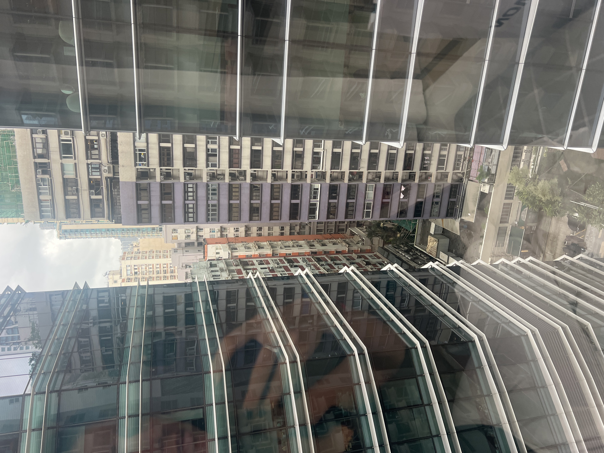 This is an outdoor, eye-level, medium shot of a dense urban environment in Hong Kong, China, taken on a cloudy day. The foreground is dominated by the reflective glass facade of a modern skyscraper, with its angled, segmented design creating a strong geometric pattern. The glass reflects other buildings, creating a mirrored effect of the cityscape.
In the middle ground and background, a cluster of older, mid-rise buildings are visible. These buildings are packed closely together and feature a variety of architectural styles and colors, including light beige, purple, and orange. They have numerous windows, some with air conditioning units protruding. The density of the buildings suggests a highly urbanized area.
The lower portion of the image shows a glimpse of a street level, with some trees and smaller structures visible between the buildings. The overall impression is one of a densely populated, vertical city with a mix of modern and older architecture.