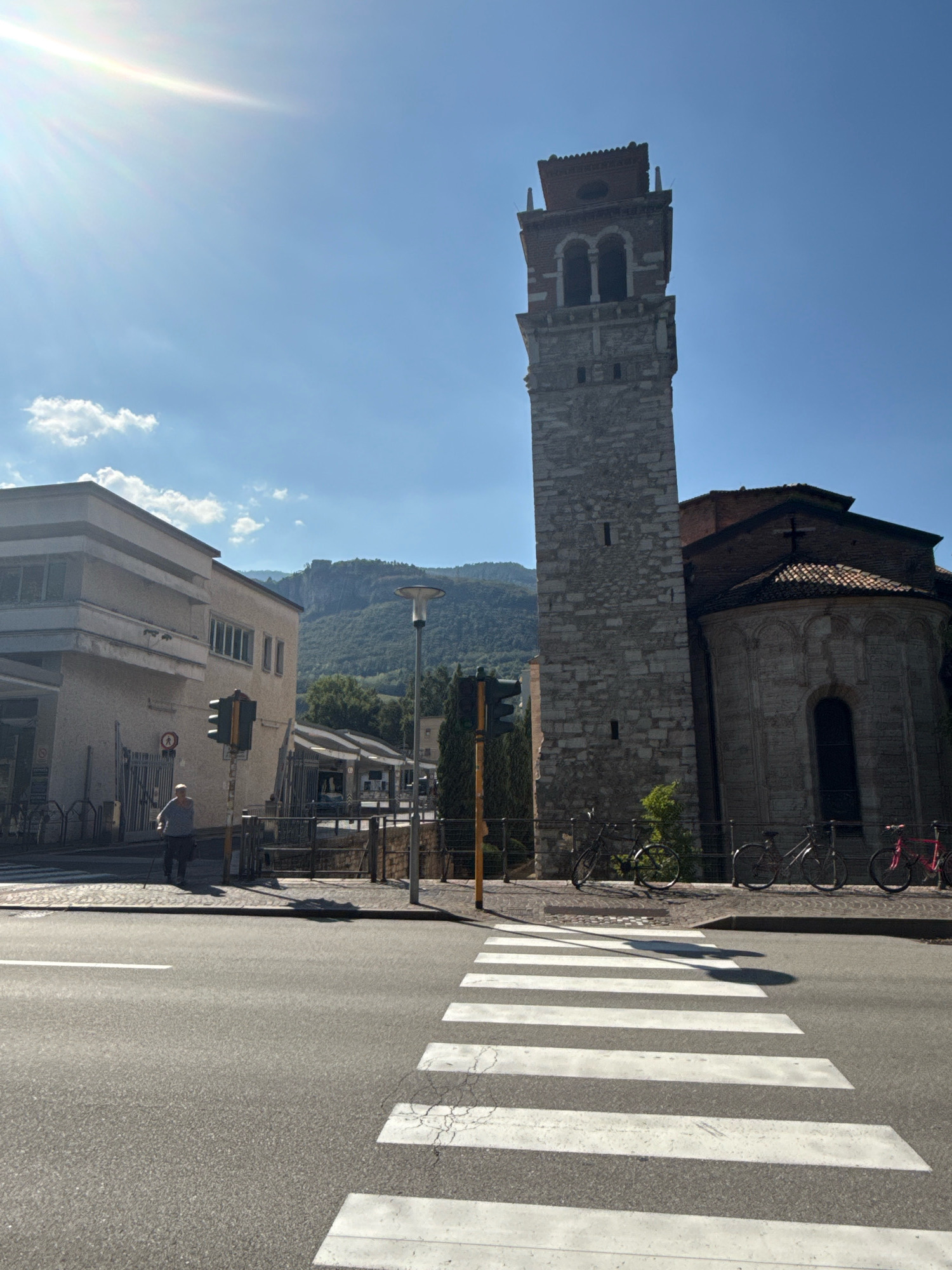 The image shows a view of a street in the city of Trento, Italy, on a sunny day. In the foreground, there is a pedestrian crossing. To the right, a tall stone bell tower with red brick accents rises against a clear blue sky. Adjacent to the tower is a church with a rounded apse and a tiled roof, featuring a simple cross. Several bicycles are parked along a railing in front of the church. To the left of the bell tower, a modern building with a light-colored facade is visible. A street light and a traffic signal are also present. In the middle ground, a person is walking with a cane on the sidewalk. In the background, lush green hills rise, suggesting a scenic mountainous landscape. The sun is shining brightly, casting shadows and creating lens flare in the upper left corner of the image.