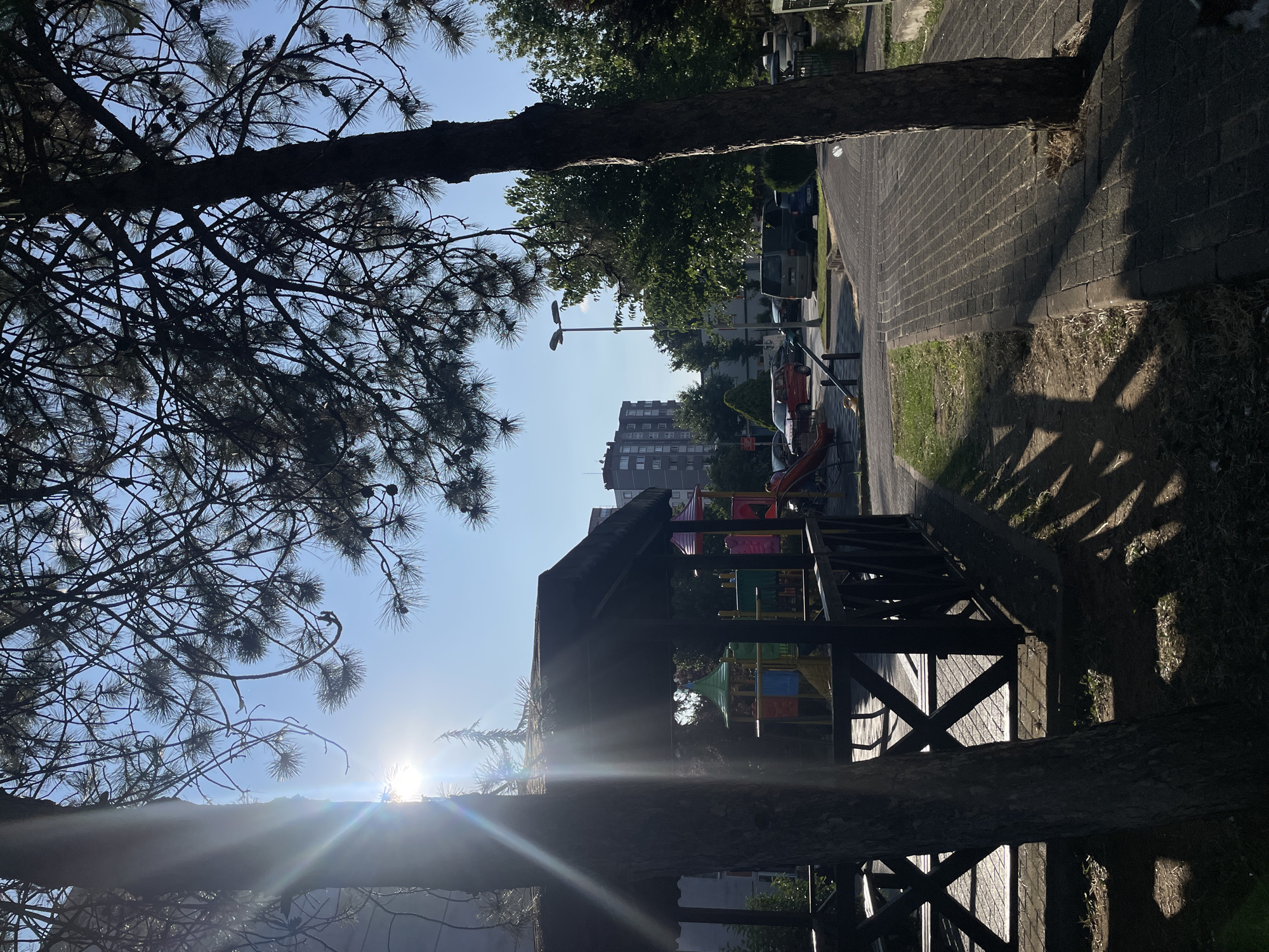 The image depicts an outdoor scene in the city of Başakşehir, Turkey, likely a residential area or park, on a clear day with bright sunlight. In the foreground, a dark wooden gazebo with a slatted roof and railings is partially obscured by the trunk of a large tree. Sunlight streams through the branches of the tree and creates sharp shadows on the ground and the gazebo's floor, which appears to be paved with brick.
Beyond the gazebo, a children's playground is visible with colorful structures, including slides and climbing frames. A pathway made of interlocking bricks runs alongside the playground and into the distance. Several cars, including a red hatchback, a silver van, and a dark blue car, are parked along the side of the road. Tall green trees and multi-story apartment buildings form the backdrop. The sky is a clear, bright blue. There are no people visible in the image.