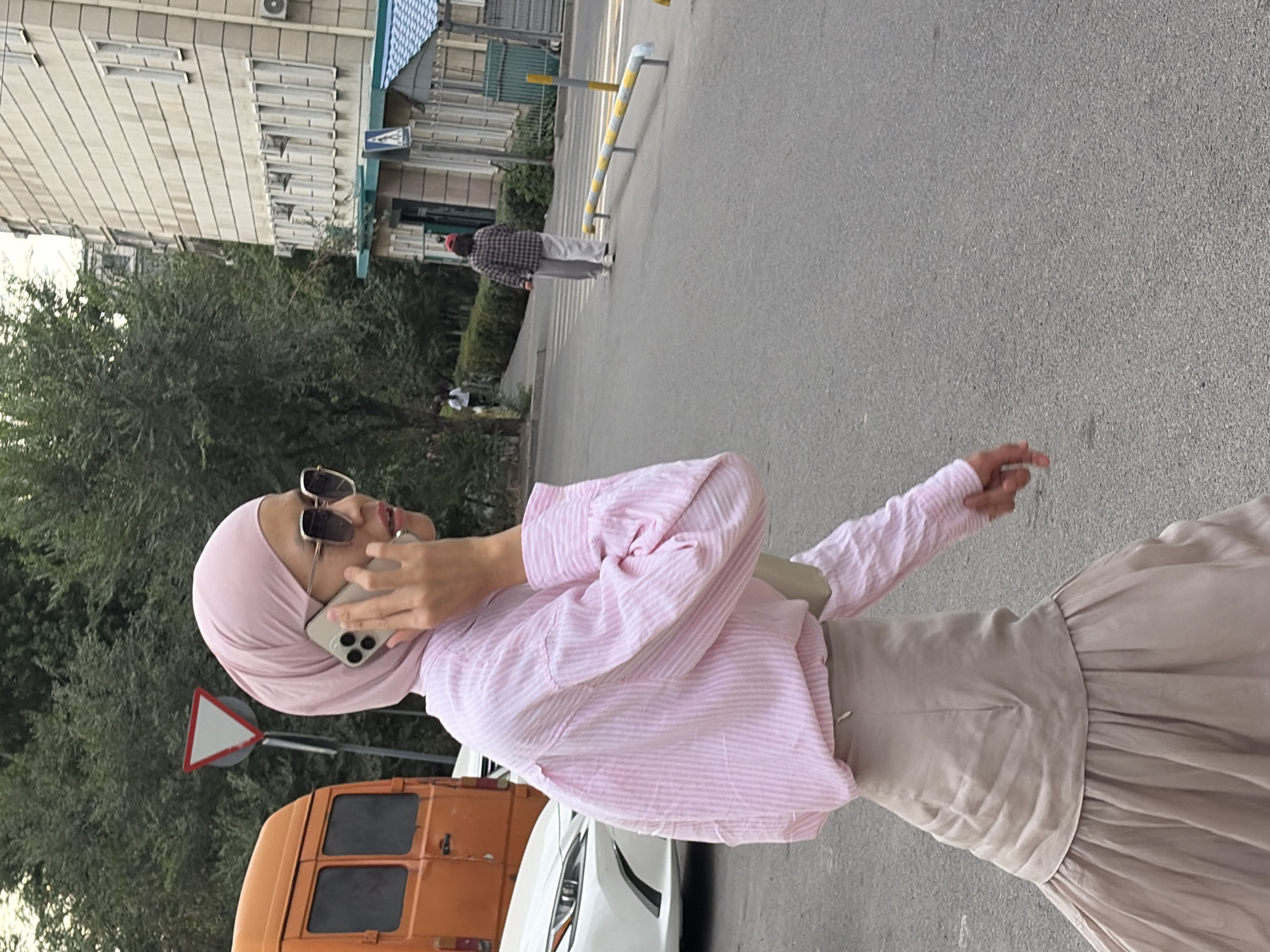 In the city of Unknown, Unknown, a woman wearing a pink hijab, sunglasses, and a pink striped button-up shirt is talking on her phone while walking. She is also wearing a beige skirt. To her left is an orange van and a white car. In the background, there is a building with many windows and trees. Another person is walking in the distance, wearing a checkered shirt and grey pants.