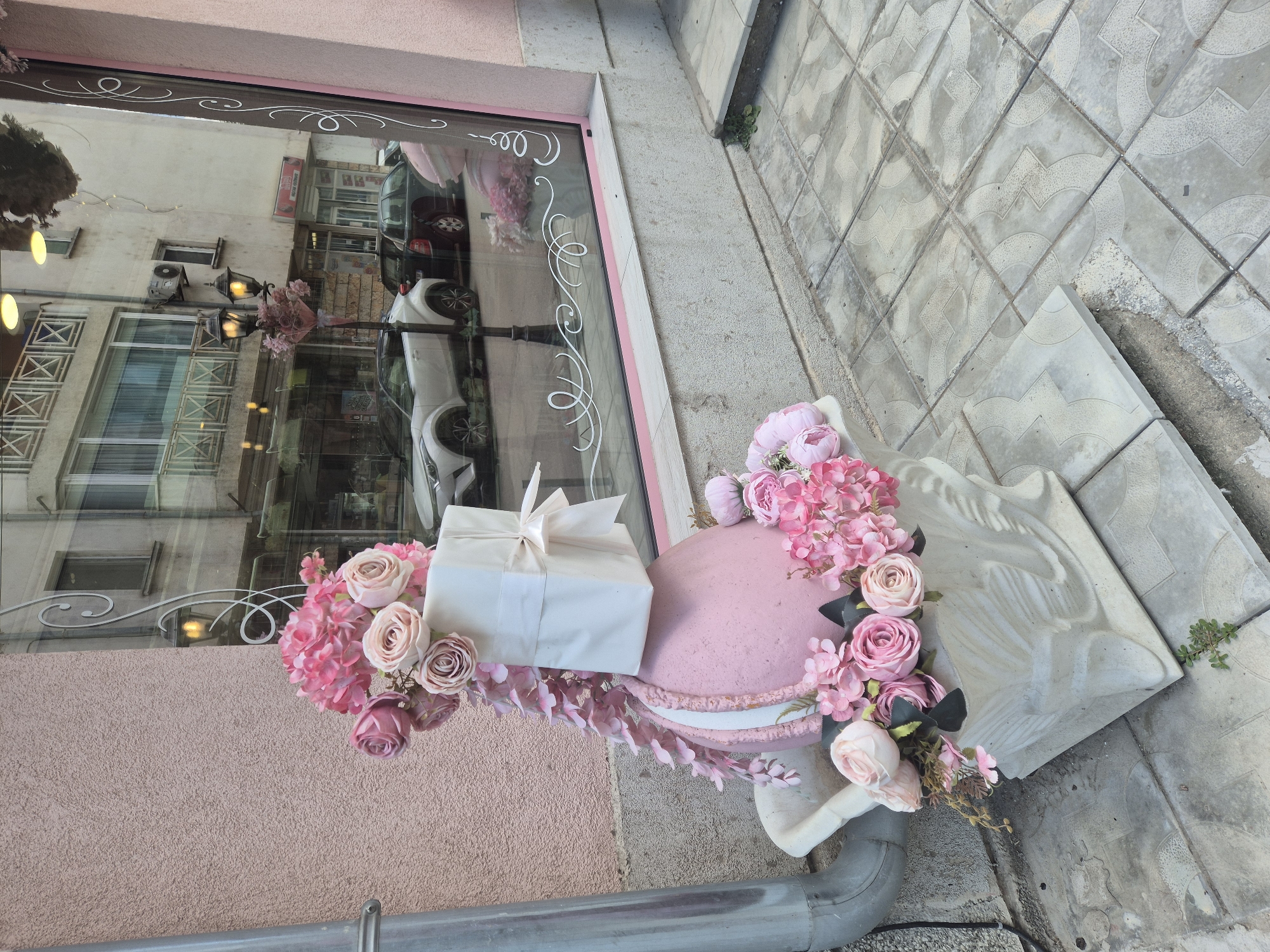 This image captures an outdoor display in front of a shop window. The primary subjects are a large, pink, macaron-shaped decoration adorned with an assortment of pink and blush artificial flowers. On top of the macaron sits a wrapped gift box with a white ribbon tied in a bow. The scene is set on a sidewalk paved with light gray interlocking tiles.
The shop window features decorative white swirl patterns on the glass. Reflections in the window show buildings across the street, including cars and shop fronts, suggesting an urban or commercial setting. One reflection clearly shows a white car, and another shows a dark-colored car. There is also a streetlamp visible in the reflection, partially obscured by a bouquet of pink flowers.
The overall impression is one of a festive or celebratory decoration, possibly for an event like a birthday or anniversary, or as part of a shop's window display. The lighting appears natural, suggesting it might be daytime, though the exact time is difficult to determine from the image. The weather seems pleasant, with no rain or harsh sunlight visible. The emotions evoked are likely positive, conveying a sense of sweetness and charm. There is no visible text in the image, except for what appears to be text on shop signs reflected in the window, which are not legible. A gray drainpipe is visible in the lower portion of the image, grounding the scene to its street-level context.