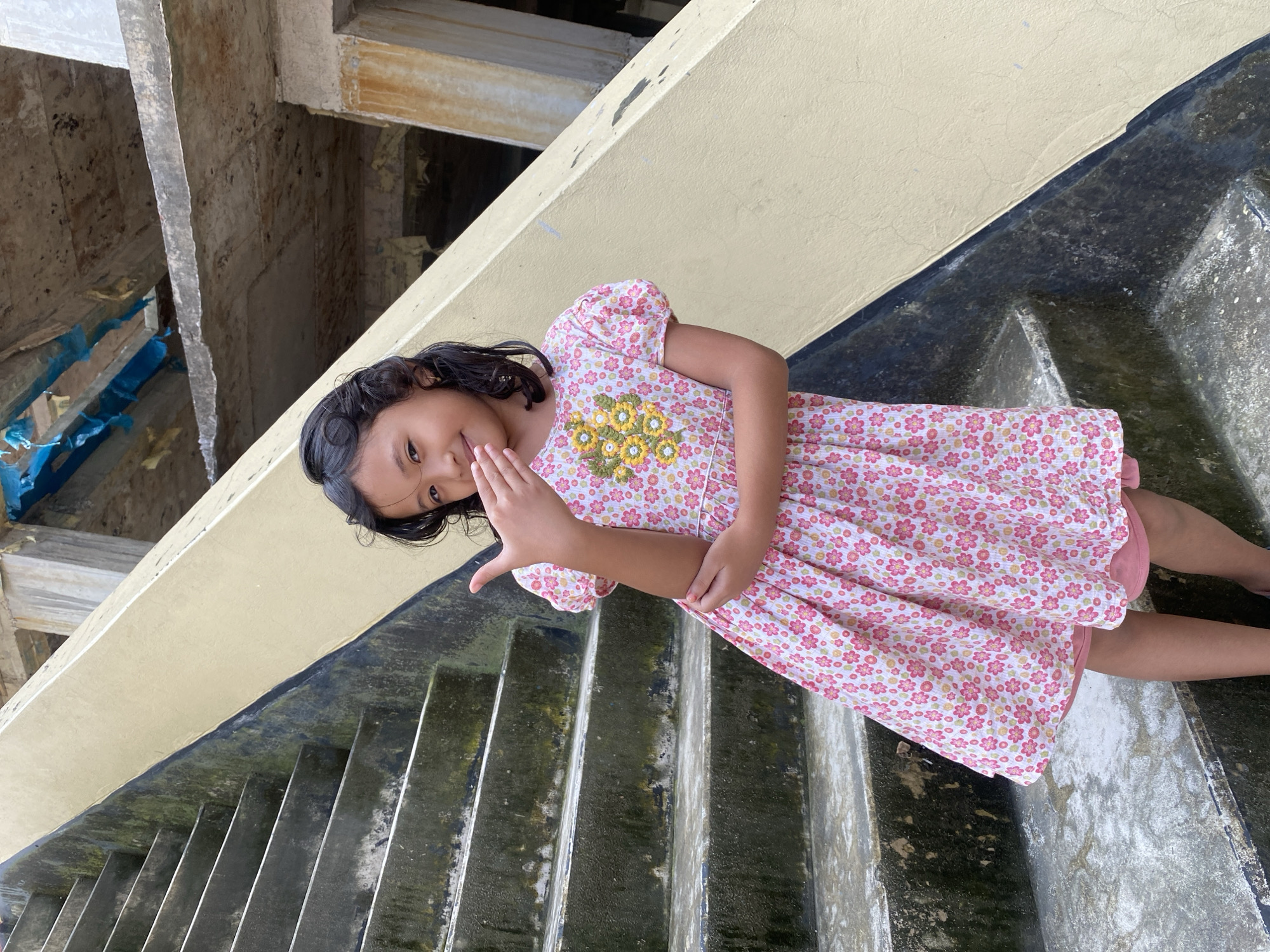 A young girl with dark, curly hair stands on a concrete staircase, partially obscured by her hand covering her mouth. She wears a pink floral dress with short, puffy sleeves and a circular yellow floral applique on the chest. The stairs are weathered and appear to be outdoors, with a cream-colored wall on the right. Above the wall, parts of an unfinished concrete structure are visible, with blue plastic sheeting. The setting is likely an urban area in Dhaka, Bangladesh, possibly an under-construction site or an older, neglected building.