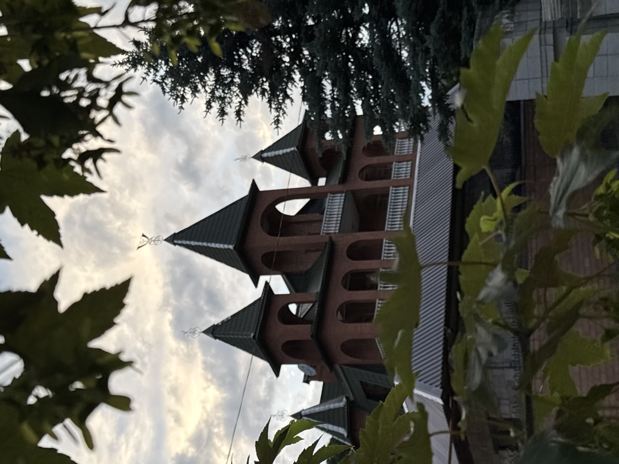 The image depicts a partially obscured view of a brick building with multiple turrets and a dark green roof, situated in Karachayevsk, Russia. The architecture suggests a style that might be described as neo-Gothic or inspired by castle-like structures. The building is framed by out-of-focus green leaves in the foreground, suggesting a naturalistic or garden setting. A tall, dark green evergreen tree stands to the right of the building, partially obscuring it and adding to the sense of depth. The sky above is overcast with dramatic clouds, hinting at either dawn or dusk. The overall impression is one of a grand, somewhat imposing structure nestled within a natural landscape, under a moody sky. There are no people or animals visible in the frame, and no actions or interactions are taking place. The scene appears to be a still, observational shot of the building and its surroundings.