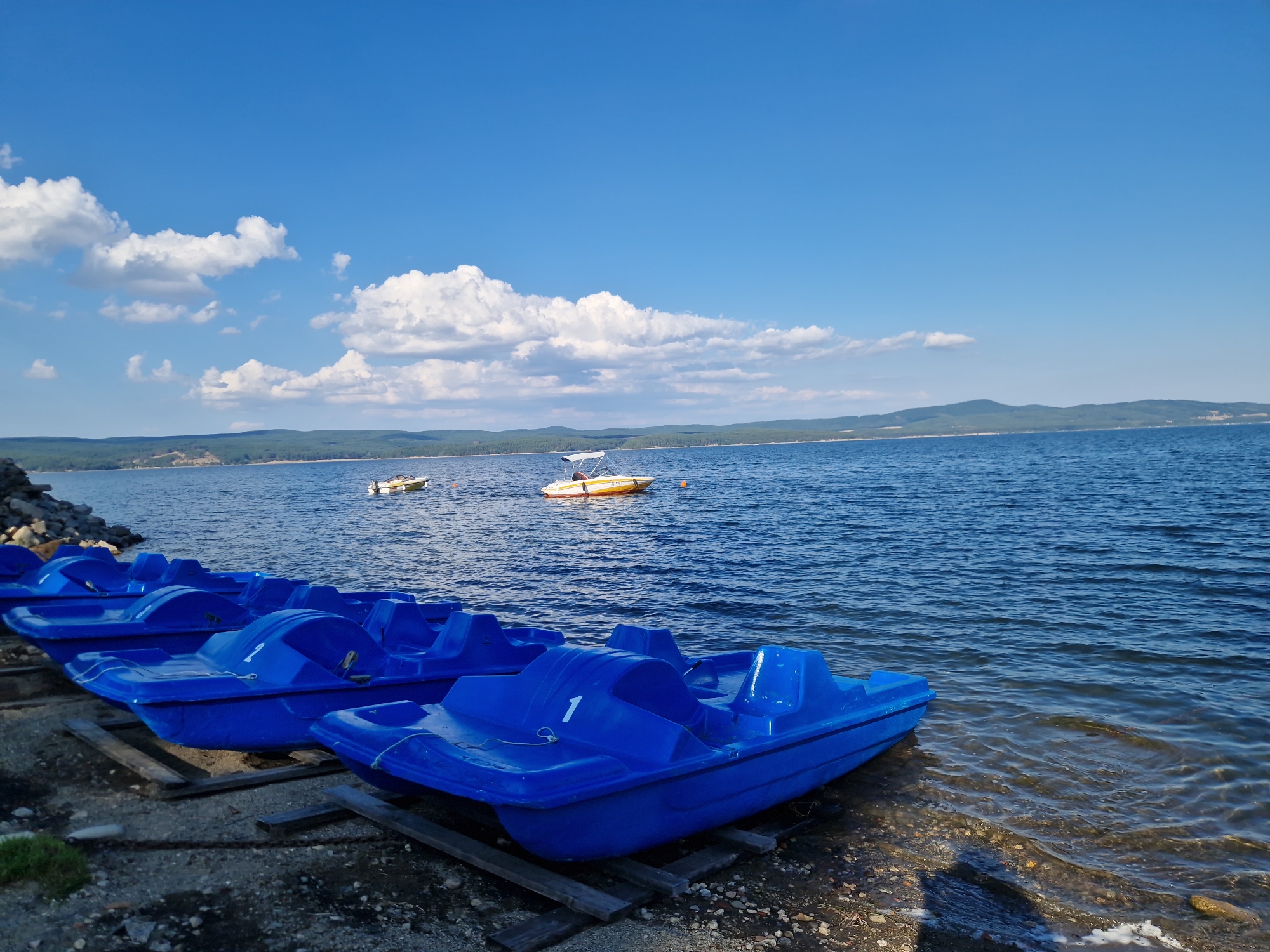 The image depicts a scenic view of the Iskar Dam in Dolni Pasarel, Bulgaria, on a clear, sunny day. In the foreground, a row of bright blue pedal boats are neatly lined up on wooden supports along the pebbled shoreline. Some of these boats have numbers visible, such as "1" on one of the closest boats. The water of the dam is a deep blue, with gentle ripples indicating a calm surface. Further out in the water, a small yellow motorboat with a white canopy is visible, and another, smaller white boat can be seen in the distance. The background features rolling hills covered in dark green trees, with a clear blue sky overhead dotted with fluffy white clouds. The overall impression is one of a peaceful and inviting recreational area.