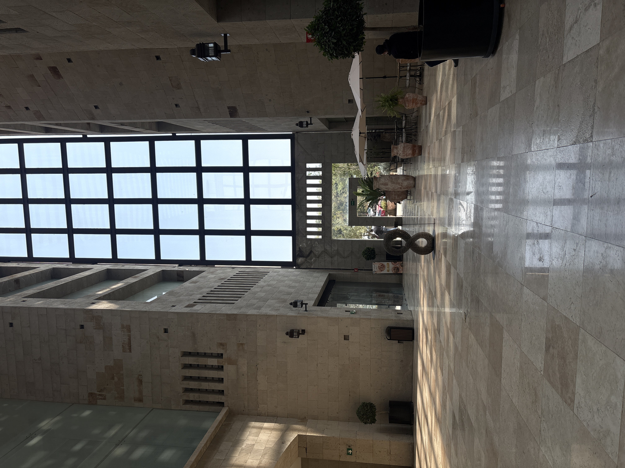The image shows an interior shot of a building in Ciudad López Mateos, Mexico. The architecture features a high ceiling with a large grid of windows that allow natural light to illuminate the interior. The floor is tiled and reflects some of the light.
To the right, there are potted plants and seating under white umbrellas, suggesting a cafe or lounge area. A person is seated at one of the tables. An art piece resembling a figure eight is visible in the foreground.
The walls are made of light-colored stone blocks with recessed designs. Security cameras are mounted on the upper portion of the walls. A sign is visible near the entrance to a store or restaurant.