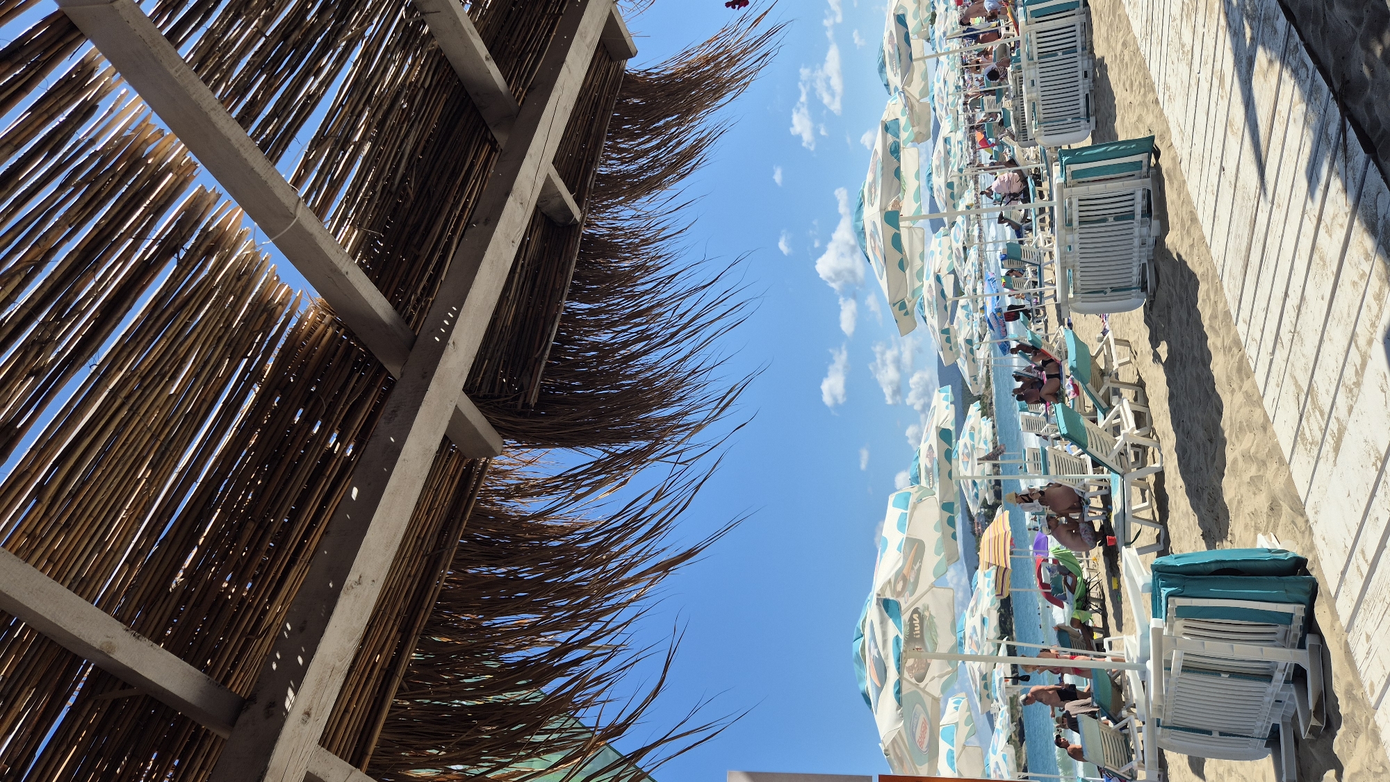The image depicts a crowded beach in Kiten, Bulgaria, on a sunny day. The foreground is dominated by a wooden boardwalk and rows of empty beach chairs and folded umbrellas, suggesting the early morning or a break in activity. Overhead, a thatched roof provides shade. Beyond this, numerous beach umbrellas, some with a white and blue geometric pattern, are set up along the sandy expanse. People are visible relaxing on lounge chairs under the umbrellas, facing the sea. In the background, the turquoise waters of the Black Sea meet a clear blue sky with scattered white clouds. The overall scene conveys a typical summer day at a popular seaside resort.