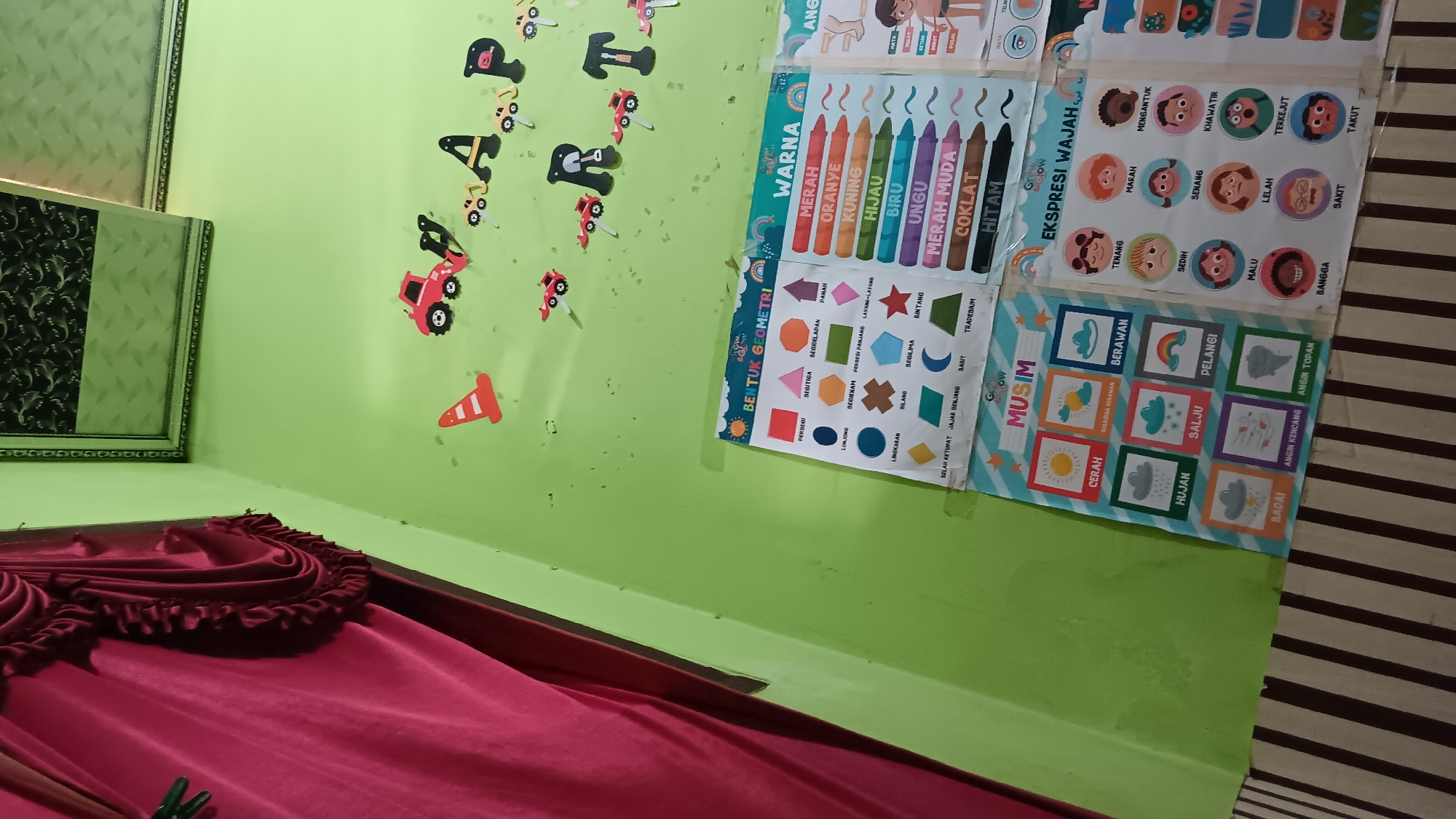 The image shows a wall with various educational posters and decorations, likely in a classroom or child's room. The wall is painted light green. A red curtain is visible on the left side of the image.
On the wall, there are several cutouts of red toy cars and traffic cones. A banner made of letter cutouts spells out "RART".
Below the banner are three educational posters:
1. A poster titled "BENTUK GEOMETRI" (Geometric Shapes) displays various geometric shapes with their names in Indonesian.
2. A poster titled "WARNA" (Colors) lists several colors with crayon illustrations next to them.
3. A poster titled "MUSIM" (Seasons/Weather) shows illustrations of different weather conditions like sunny, cloudy, rainy, snowy, rainbow, stormy, windy, and tornado, along with their Indonesian names.
4. A poster titled "EKSPRESI WAJAH" (Facial Expressions) displays cartoon faces depicting different emotions such as calm, angry, surprised, sad, happy, worried, shy, tired, proud, sick, and scared, with their Indonesian names.
The floor is partially visible at the bottom, showing a striped pattern of brown and off-white. The image's perspective suggests it was taken from a low angle, looking up towards the wall. The lighting appears to be artificial, possibly from an overhead source.
This scene is located in the city of Sonobekel, Indonesia.