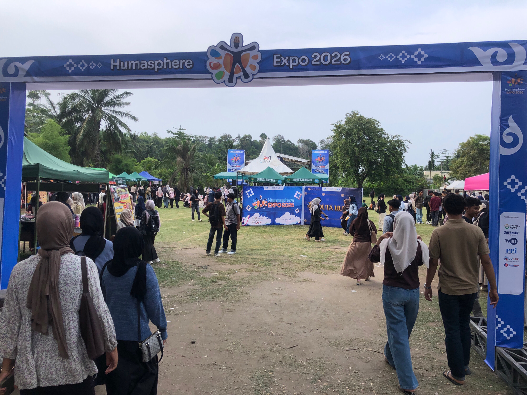 The image depicts an outdoor event, likely an expo, at Universitas Malikussaleh in Lhokseumawe, Indonesia. A large blue banner arching over an entrance reads "Humasphere Expo 2026" with a logo of stylized human figures. Various tents and booths are set up in the background, some with banners displaying "Universitas Malikussaleh" and "EXPO 2026". A crowd of people, many wearing headscarves and casual attire, are milling around the grounds, walking and standing. The ground is a mixture of dirt and grass, and palm trees and other greenery are visible in the background under a cloudy sky. Several people are walking away from the camera towards the expo area, while others are gathered near the entrance. A blue structure with logos and text, potentially a sponsor's display, is on the right side of the frame. The overall atmosphere appears to be that of a community event or academic fair.