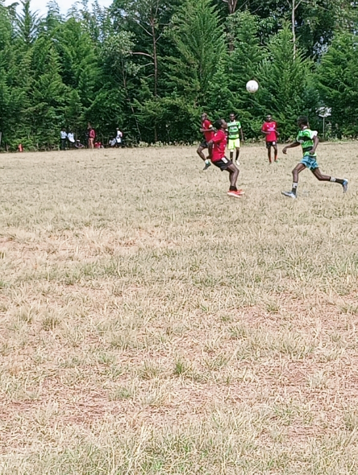 An outdoor football match is in progress on a dry, grassy field in Lwandeti ward, Kenya. A male player wearing a red jersey, dark shorts, and orange footwear is captured mid-air, having just kicked a white football with his right foot. The football is visible above and slightly to his right. Another male player in a green and black striped jersey, patterned shorts, and dark footwear is running towards the right in the mid-ground. Additional players in red and green/black jerseys are positioned across the field, indicating active play between two teams. A dense line of tall green trees forms the background, with several indistinct individuals visible along the tree line to the left. The field itself consists of light brown and green sparse grass with visible patches of earth.