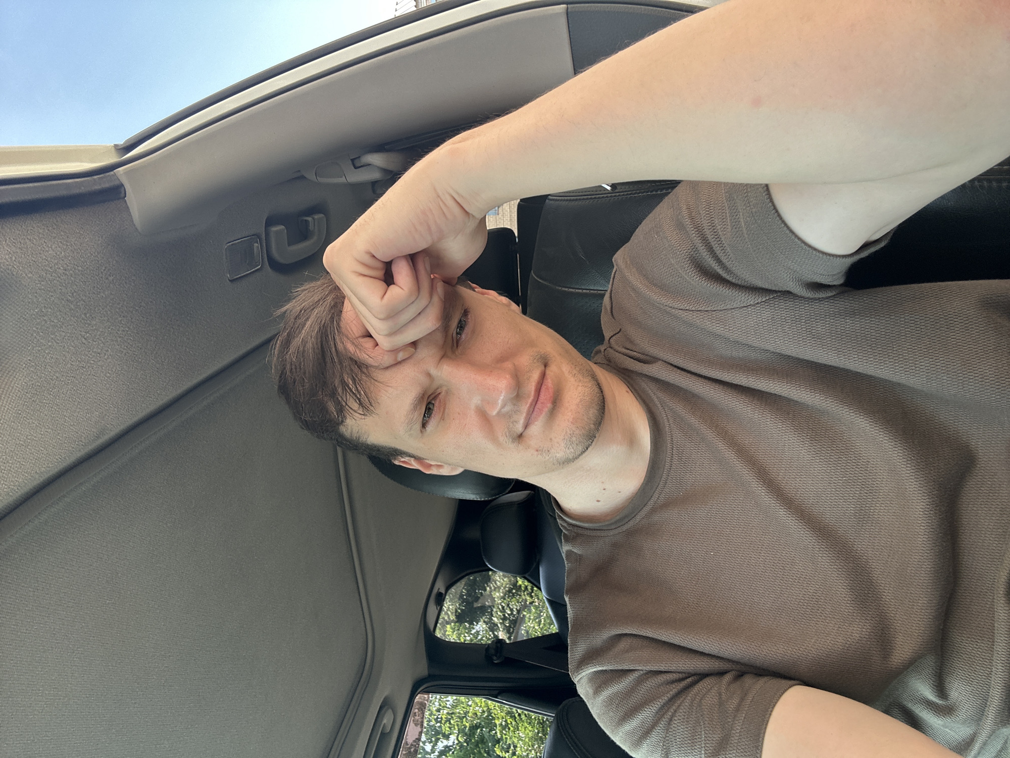 A selfie-style, medium shot captures a man sitting in the driver's seat of a car, with a sunroof overhead and a clear blue sky visible through the passenger side window. He is wearing a brown t-shirt and has his right hand on his forehead, with his gaze directed towards the camera. The interior of the car appears to be dark leather, with the headrest of the passenger seat visible behind him. The background outside the window shows lush green trees and foliage. The man has short, brown hair and appears to be in his late 20s or early 30s. His expression is a mix of weariness and a slight smirk. The image is taken from a slightly elevated angle, looking down at the man. The location in the city of Shatava, Ukraine, is not discernible from the image itself.