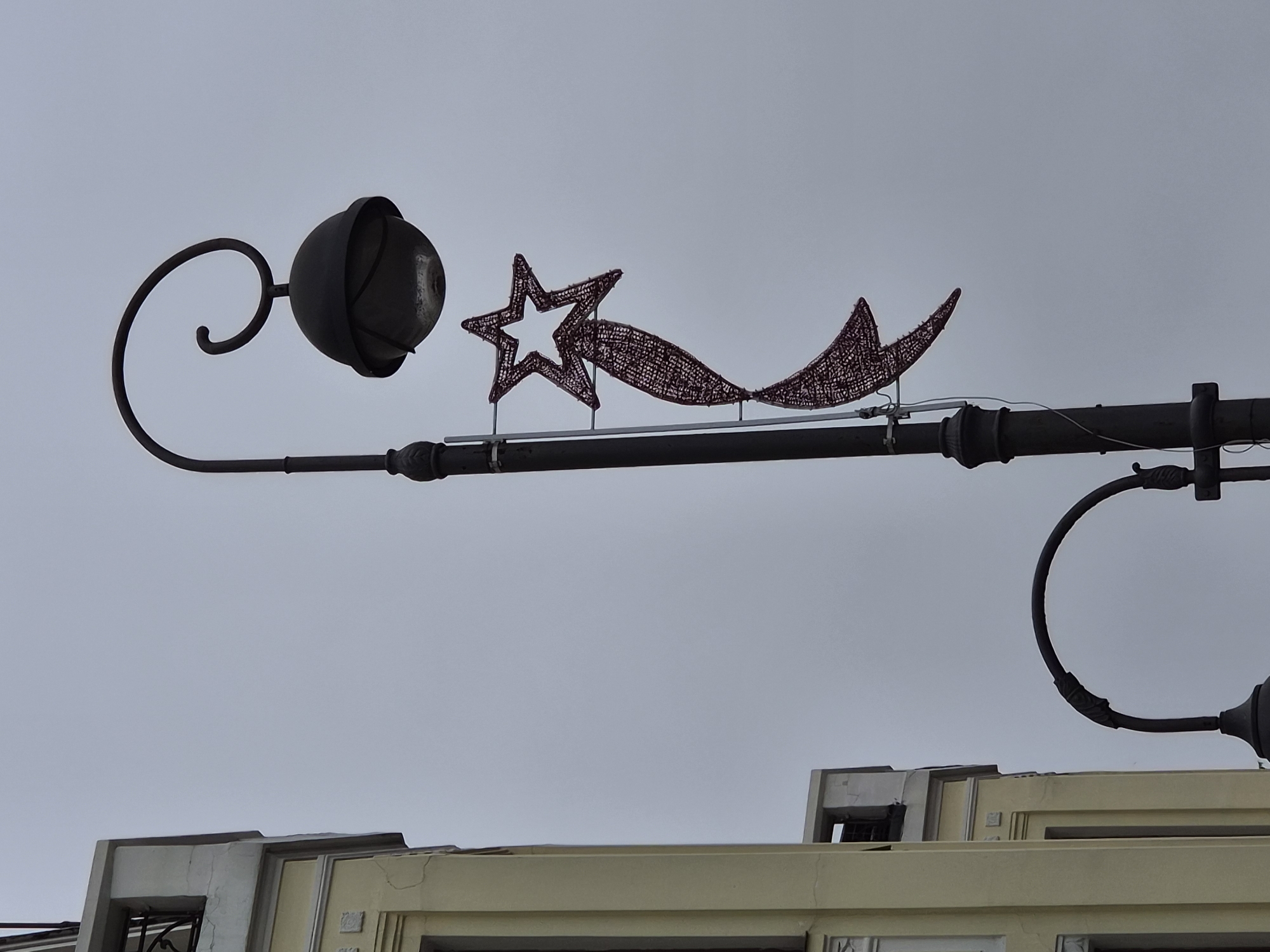 The image shows a street lamp with a Christmas decoration resembling a shooting star. The lamp post is dark and ornate, with a curved arm holding a spherical lamp. The shooting star decoration is made of red, mesh-like material and consists of a star shape with a trailing tail. To the left of the lamp post, a section of a pale yellow building with decorative architectural details is visible. The sky is overcast and gray. In the foreground and partially obscured by the building, another street lamp with a curved arm is visible. The scene appears to be in an urban environment. The specific location is the entrance to the "Dom Druzhby" (House of Friendship) on Svetlanskaya Street, Vladivostok, Russia.
