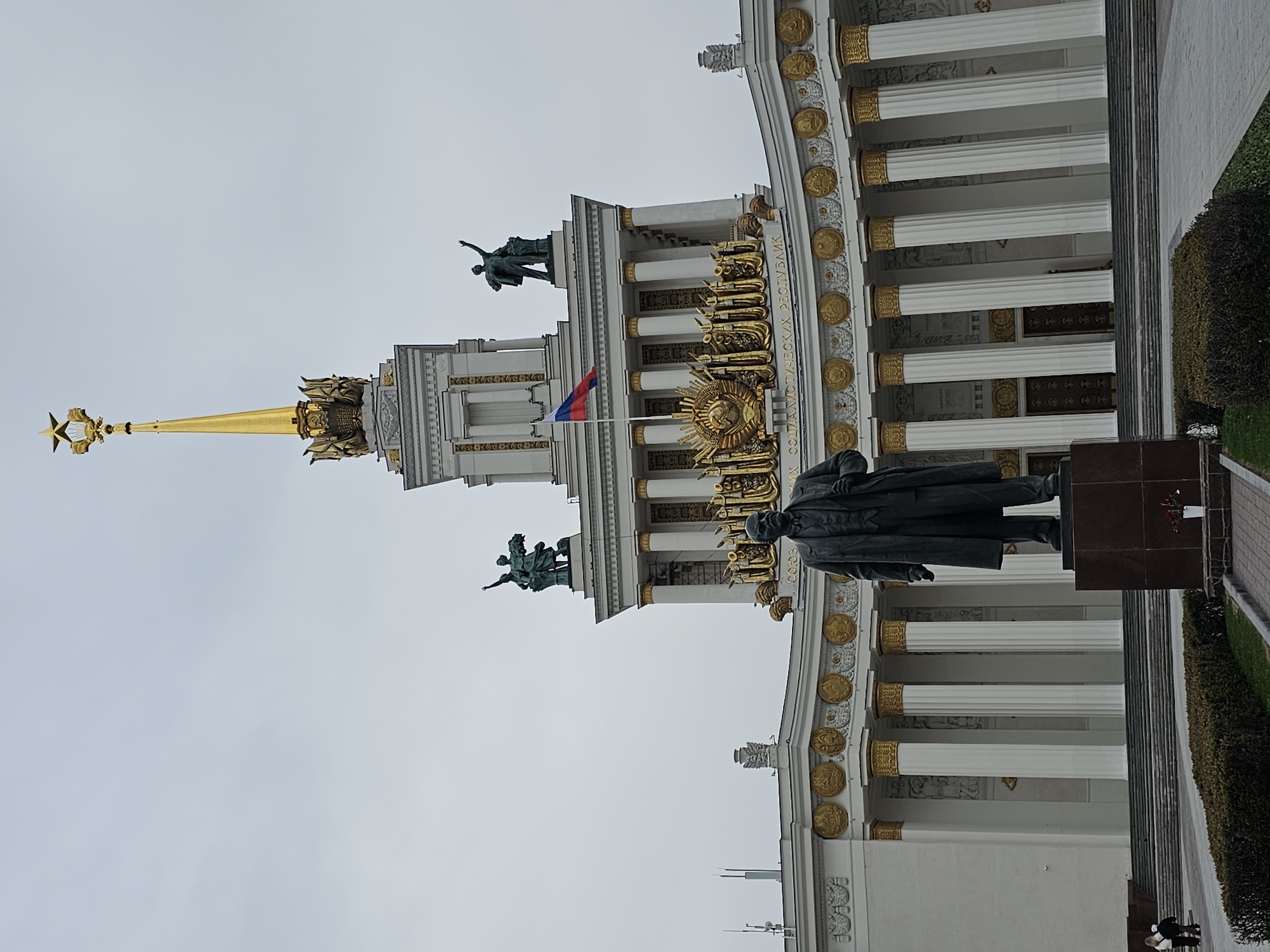 The image displays a grand, ornate white building featuring extensive golden ornamentation under an overcast sky. A tall, golden spire topped with a large golden five-pointed star crowns the central section of the building. On the roofline, statues of human figures, possibly allegorical, are visible on multiple levels. A Russian flag flies from a pole on the building's facade.
In the foreground, a dark-colored statue of Vladimir Lenin stands on a brown pedestal, facing the building. The building's architecture is characterized by numerous white columns with golden capitals and bases. Its facade is richly decorated with golden medallions, wreaths, and Soviet-era symbols, including the hammer and sickle. Clearly visible text in gold letters on the facade spells out "СОЮЗ СОВЕТСКИХ СОЦИАЛИСТИЧЕСКИХ РЕСПУБЛИК" (Union of Soviet Socialist Republics).
Manicured green bushes line a paved area to the right of the Lenin statue. The scene suggests a monumental public space, likely from the Soviet period given the architectural style and symbols, located in Moscow, Russia. The time of day is daytime, but the uniformly gray sky indicates overcast weather. No specific activity or event is taking place; the scene is static. A few indistinct figures are barely visible in the far background near the bushes.