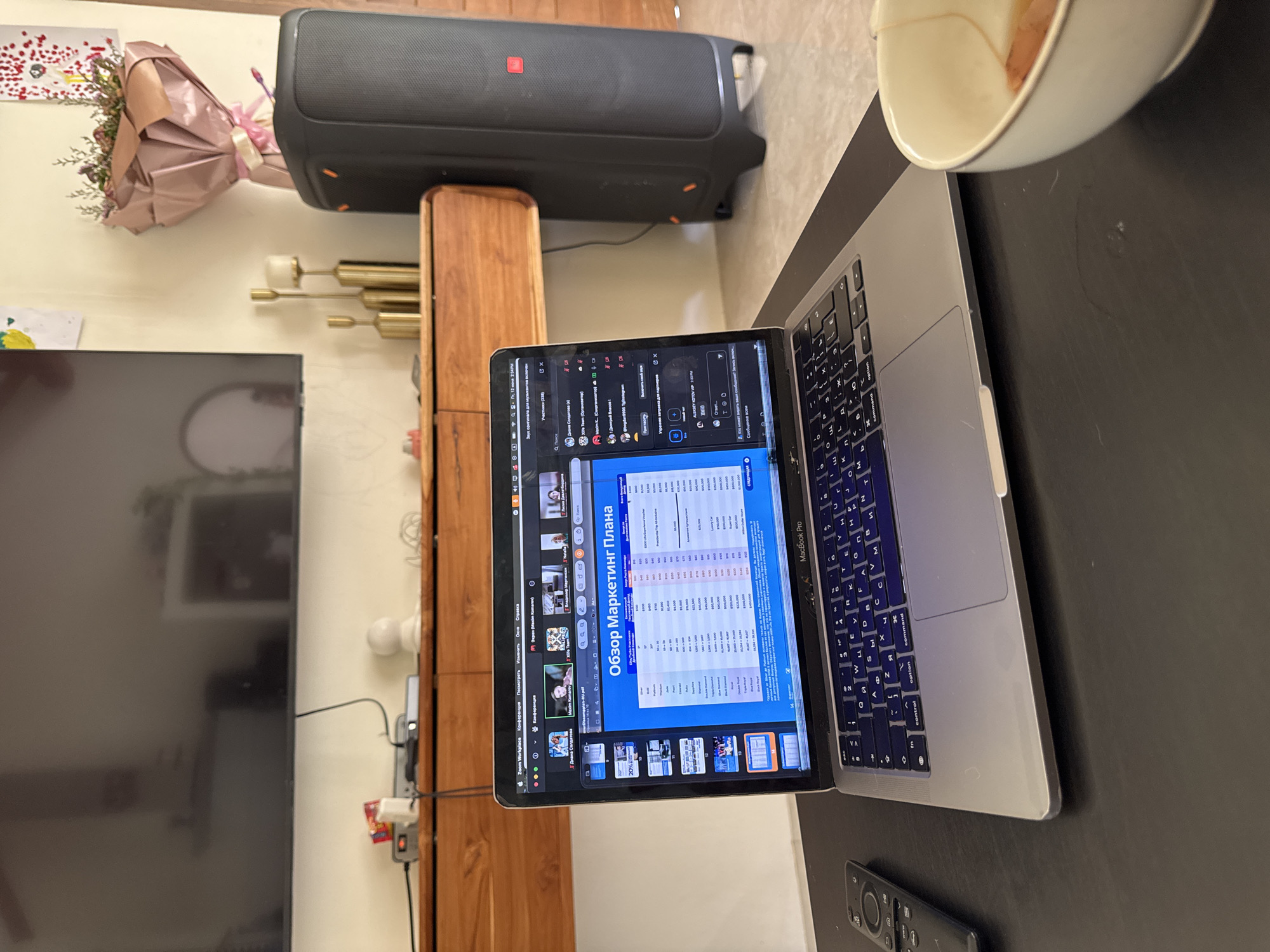 The image shows a MacBook Pro open on a dark surface. The laptop screen displays a presentation with Russian text, titled "Обзор Маркетинг Плана" (Marketing Plan Overview). Several participants are visible in video calls on the screen. To the left of the laptop is a black remote control. In the background, a large black speaker system stands to the right of a wooden cabinet. A bouquet of flowers wrapped in pink paper is placed on top of the speaker. Above the cabinet, a large dark television screen is mounted on the wall. The environment appears to be indoors, possibly a home or office setting. The location is specified as ตำบลเกาะพะงัน, Thailand, however, there are no visual cues in the image to confirm this specific location.