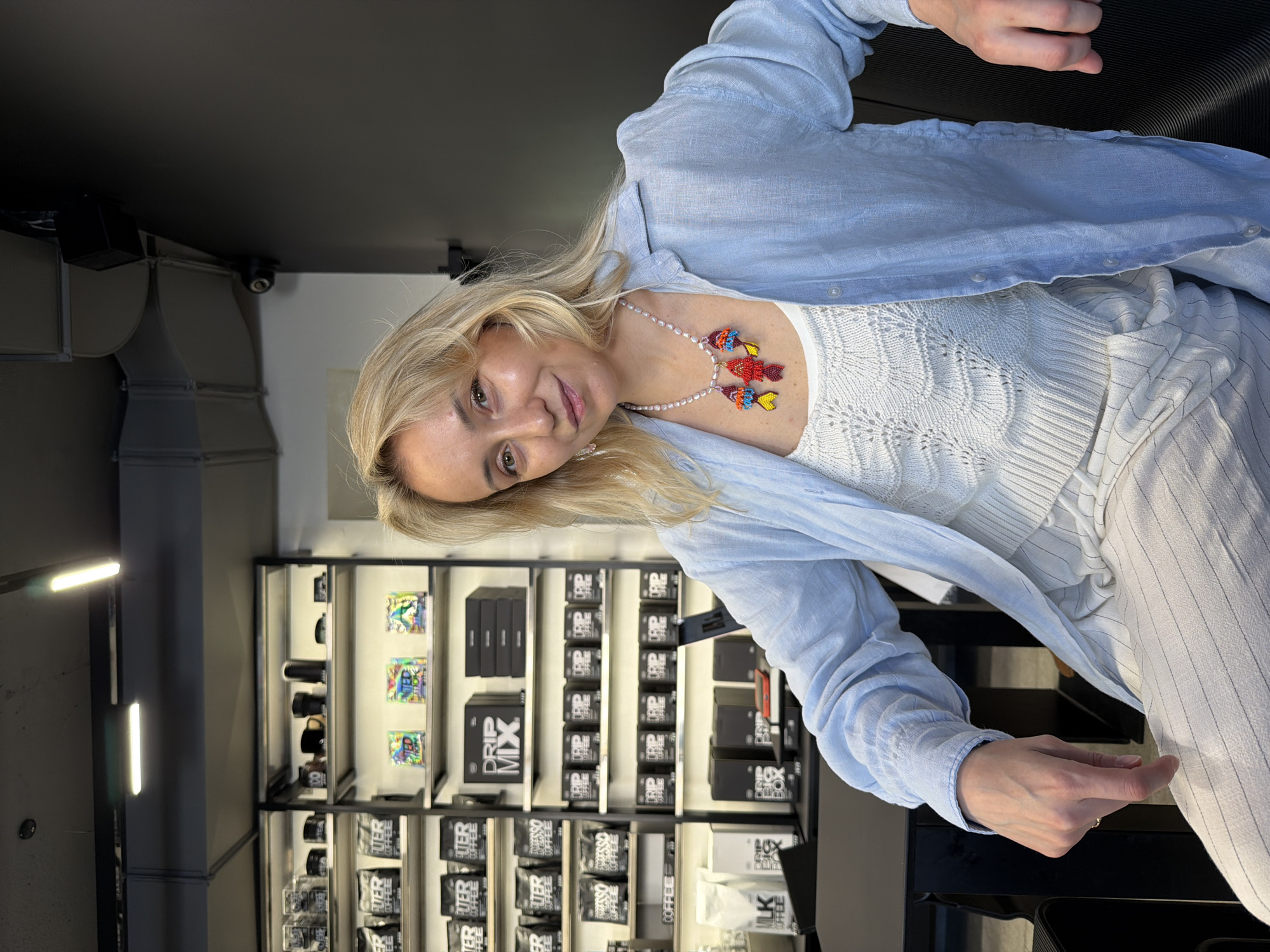 The image depicts a blonde woman with fair skin, wearing a light blue button-down shirt over a white, textured top, and striped white pants. She is also wearing a pearl necklace with colorful beaded fish pendants. The woman is sitting indoors, with her head tilted slightly towards the camera.
Behind her, a retail display is visible, featuring shelves stocked with various coffee products. Some of the items include black boxes labeled "DRIP MIX" and "DRIP COFFEE", as well as bags of coffee labeled "SWISSO COFFEE".
The location appears to be a retail space, possibly a coffee shop, in Izhevsk, Russia.