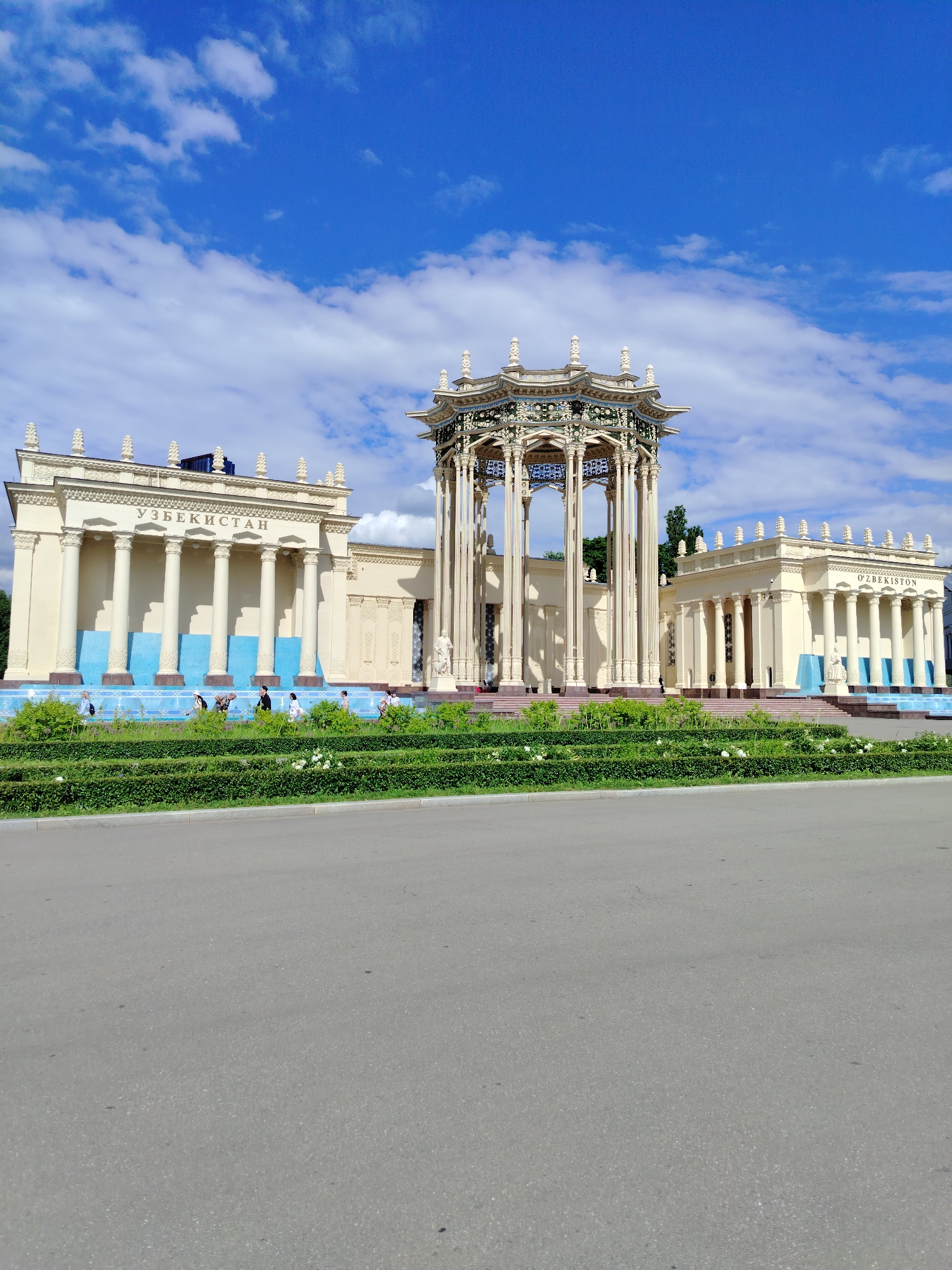 This is a vertical, medium-shot photograph of the Uzbekistan pavilion at the VDNKh park in Moscow, Russia. The pavilion is a grand, classical-style building with a facade of cream-colored columns and ornate architectural details. In front of the pavilion, a vibrant blue fountain runs the width of the structure, with lush green hedges and manicured bushes bordering it. Several people are walking along the path in front of the fountain, enjoying the sunny day.
The sky above is a brilliant blue, dotted with fluffy white clouds. The bright sunlight casts shadows, suggesting it is midday. The Uzbekistan pavilion, with its distinctive name written in Cyrillic script across its facade, is a prominent landmark at VDNKh. The pavilion's intricate design, featuring a rotunda with multiple columns and decorative elements, stands out against the clear sky. The overall mood of the image is one of pleasant outdoor activity and appreciation for architectural beauty. The presence of the road in the foreground suggests a public space designed for visitors.