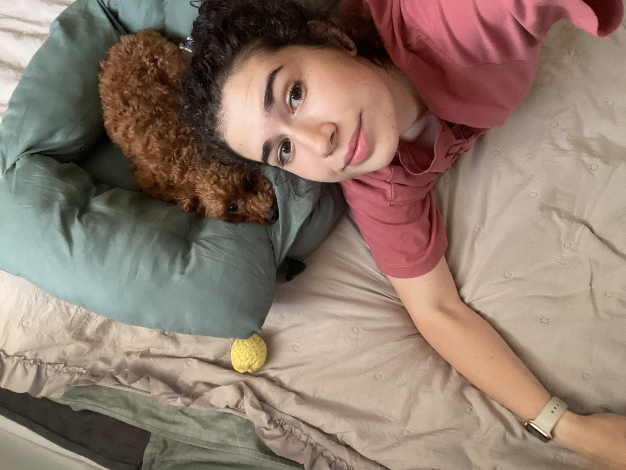A medium shot shows a woman and a small, brown poodle lying on a bed with pale beige sheets. The woman, with dark, curly hair and light skin, wears a rose-colored shirt. She is lying on her back, with her head close to the dog, and has one arm extended along the bed. The poodle is lying in a green dog bed. A yellow, knitted ball lies near the dog bed on the bed sheets. The location is unknown.