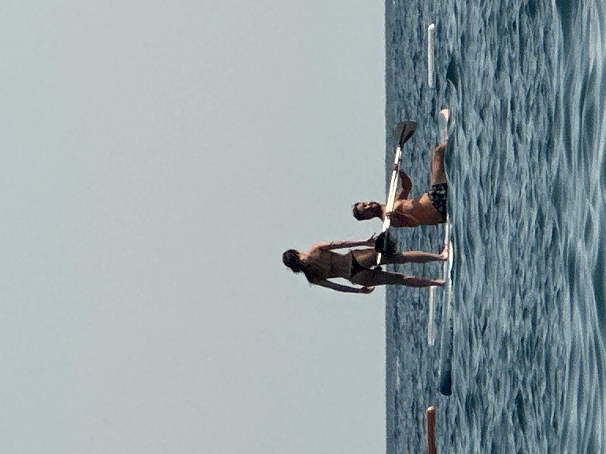 Two people are on paddleboards in the water. A woman in a bikini is standing on one paddleboard, facing away from the camera. A man is sitting on another paddleboard, holding a paddle. The water is blue and choppy, and the sky is clear and pale. The scene appears to be in the city of Boreti, Montenegro, given the presence of the Adriatic Sea.