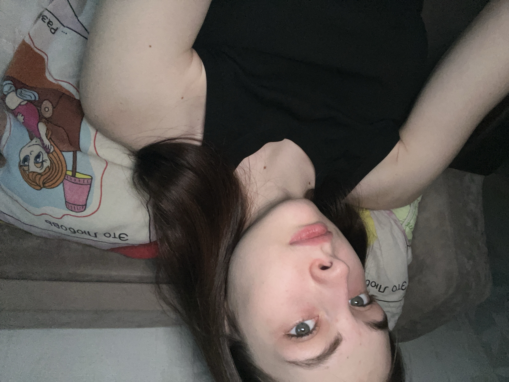 A woman is lying down, possibly on a bed, with a pillow under her head. She has dark hair and fair skin. She is wearing a black top. The pillow has a cartoon illustration and the words "Это любовь" (This is love) written on it in Russian. The background is partially visible, showing what appears to be a wall and a sofa or bed frame. There is not enough information to identify a location in Shcheglovo, Russia.