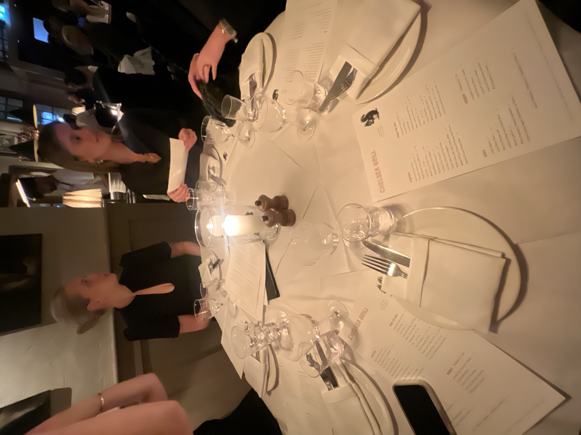 Here's a description of the image:
The image depicts a group of people seated around a round table at a restaurant. The table is covered with a white tablecloth and set with multiple plates, silverware, wine glasses, and menus. The menus are printed with the name "CHELSEA GRILL," indicating the restaurant's name.
The people at the table appear to be engaged in conversation and dining. One person is holding a white paper (likely a menu or wine list). A lit candle is present in the center of the table. The dining area seems dimly lit, indicating an evening setting.
The "Chelsea Grill" menu hints at a possible location in Chelsea, a district in London, United Kingdom.
