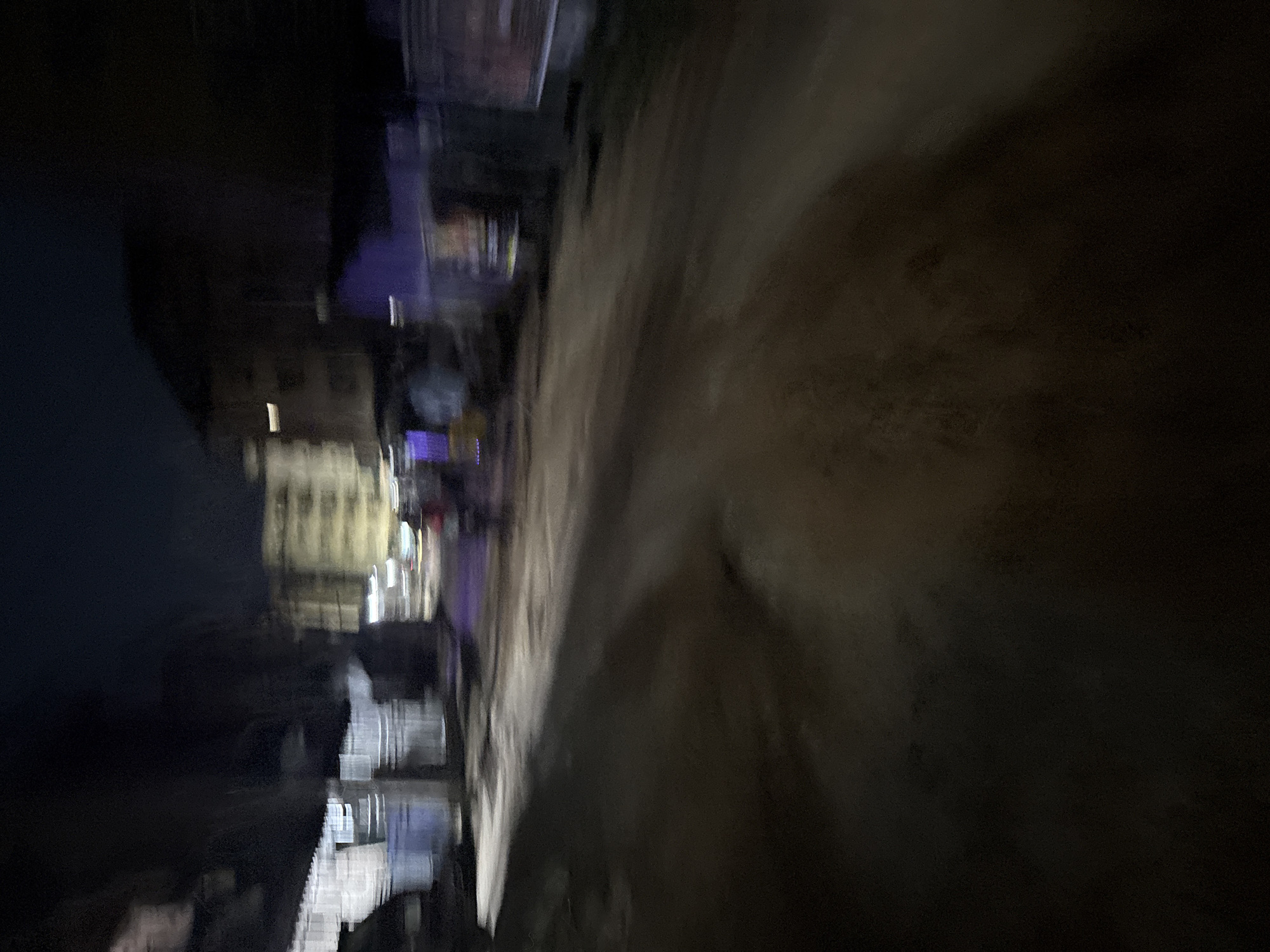 The image depicts a nighttime street scene in Onitsha, Nigeria, characterized by motion blur due to low light conditions and movement. The road surface appears to be unpaved or dirt, with some unevenness. Buildings are visible in the background, some illuminated with artificial lights, suggesting a commercial or residential area. The blur obscures specific details of the structures, but hints of multiple stories and windows can be discerned. There are indications of vehicles and possibly people in motion, appearing as streaks of light and indistinct shapes. The overall atmosphere is dark and somewhat chaotic, typical of a busy street at night in a densely populated urban environment.