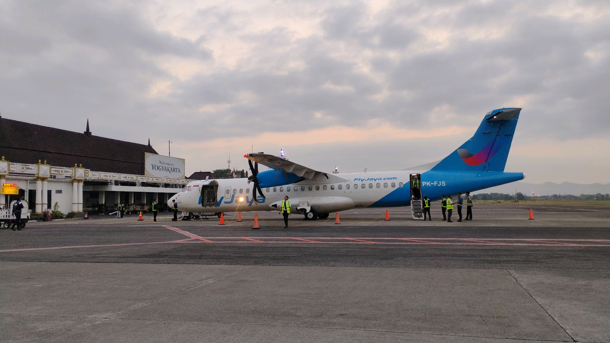 An outdoor, medium shot captures an airplane parked on an airport tarmac at dusk. The airplane, primarily white with blue accents and a blue tail with a pink and red logo, has "Fly-Jaya.com" printed on its fuselage. Several people in high-visibility vests are visible near the plane, some appearing to be airport staff and others possibly passengers. A set of stairs leads into the aircraft's cabin, which is open. Orange traffic cones are placed around the plane. In the background, a building with a dark roof and white pillars bears a sign that reads "Welcome to YOGYAKARTA." The sky is overcast with muted light. The scene suggests the operational activities of an airport, likely Adisutjipto International Airport, in the Maguwoharjo area of Yogyakarta, Indonesia.