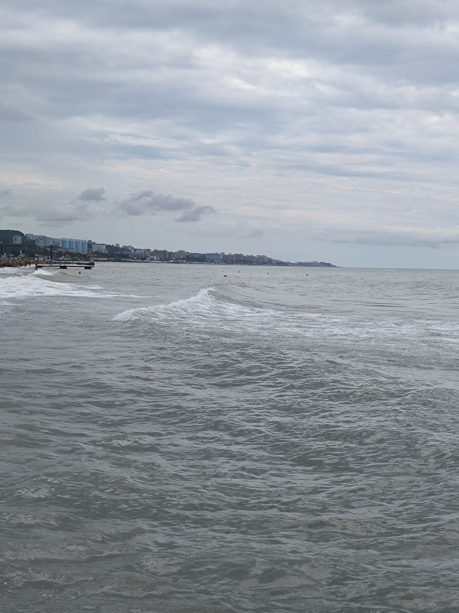 A wide-angle, eye-level shot captures the coastline of Sochi, Russia, under a heavily overcast sky. The choppy sea fills the foreground and midground, with waves breaking near a pier on the left. A distant city skyline with buildings of varying heights stretches across the horizon. The pier is populated by a few people, some of whom appear to be standing or walking. Several buoys are visible in the water. The overall impression is of a grey, somewhat bleak day by the sea.