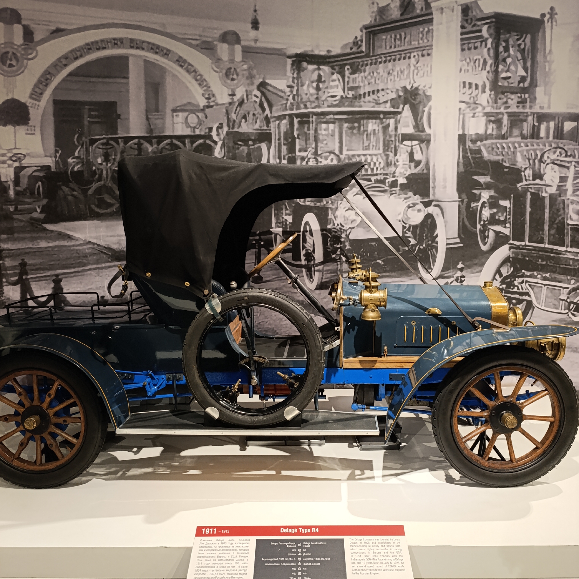 The image features a blue Delage Type R4 automobile from 1911-1913, exhibited at a museum. The car has wooden wheels, a black convertible top, and brass accents. A spare tire is mounted on the side. The background shows a black and white image of vintage automobiles and an arch with text in Russian. The scene is set on a white platform. A sign in front of the car provides details about the Delage company and the model. The image was taken at the Auto Museum in Verkhnyaya Pyshma, Russia.
