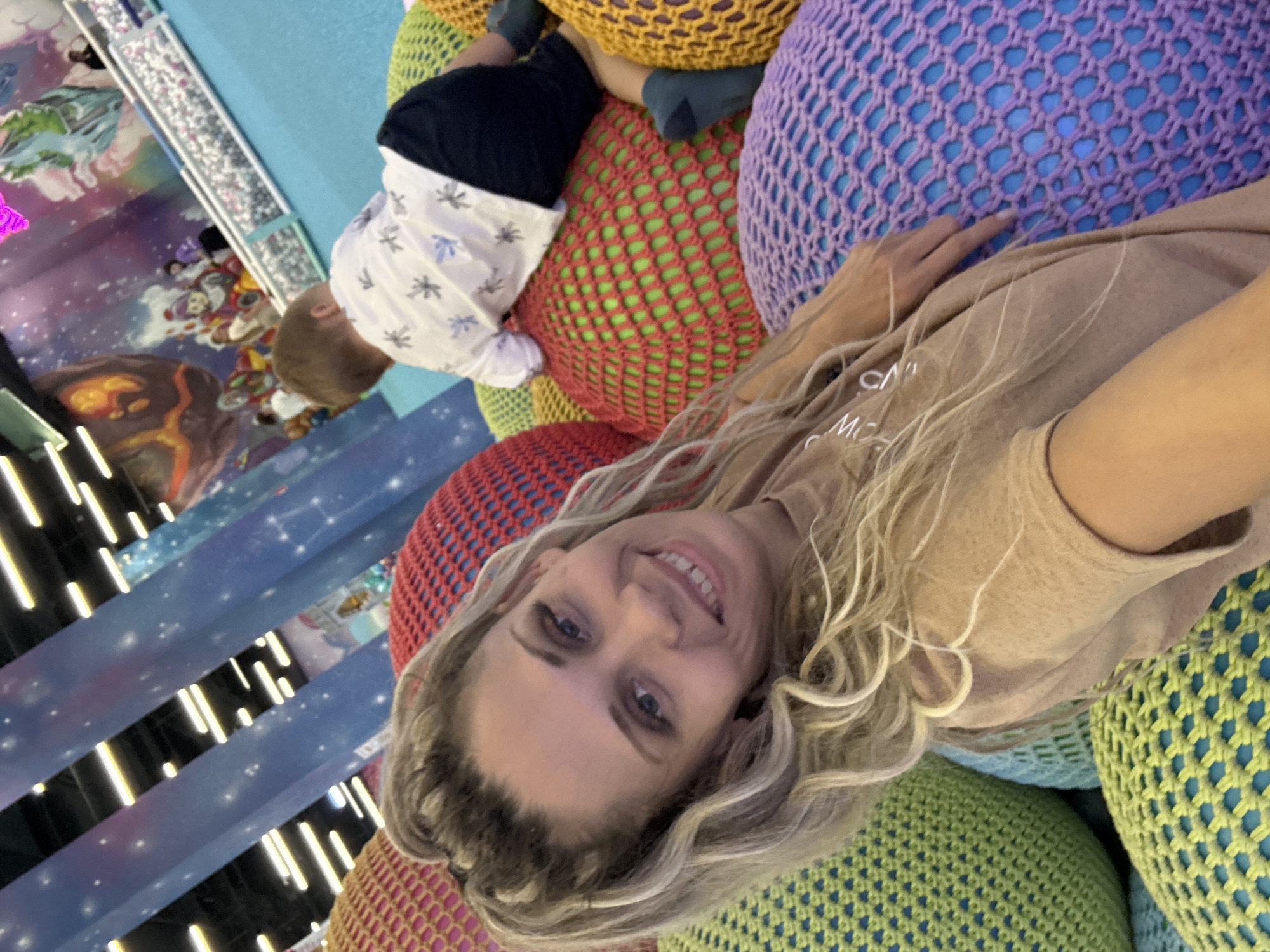 The image shows a woman and a child in what appears to be an indoor playground. The woman is in the foreground, taking a selfie, with a wide smile and blonde hair. She is wearing a tan t-shirt. Behind her, a young child is on all fours, wearing a white t-shirt with a pattern and dark shorts. They are surrounded by large, colorful, knitted poufs in shades of green, red, purple, and yellow, with blue netting underneath. In the background, there is a bright blue floor and a wall decorated with cartoonish space-themed murals, including planets and stars. There are also other people visible in the distance, as well as a ball pit. The environment suggests a playful and energetic atmosphere. The location is identified as being in the city of Zhukovo, Russia.