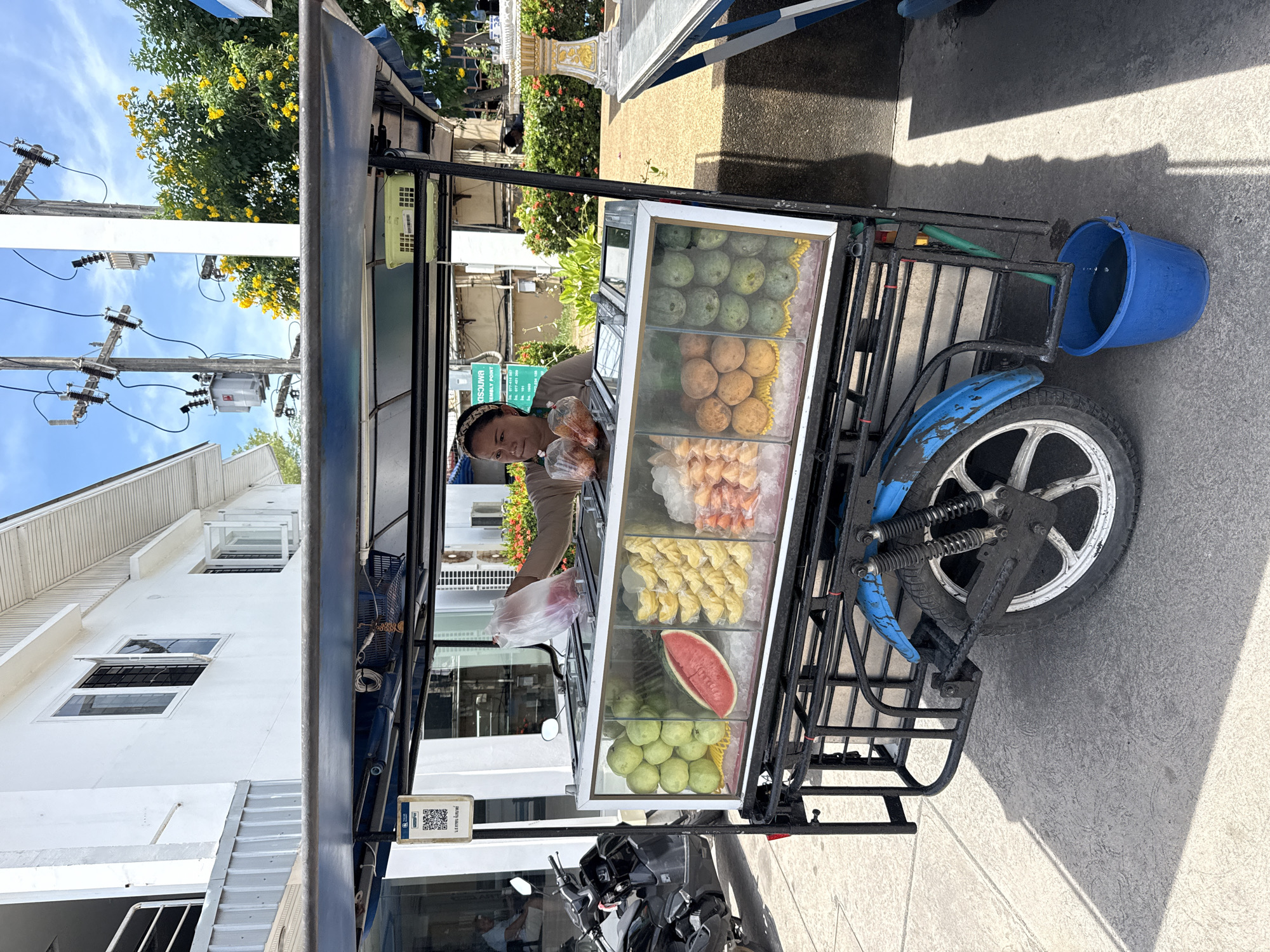 A woman stands behind a fruit cart in a city setting, likely Ko Samui District, Thailand. The cart is equipped with a glass display case filled with various fruits, including green guavas, watermelon slices, and other colorful chopped fruits. The woman is holding a plastic bag with pinkish fruit inside. Behind the cart, a white building with windows and a blue overhang is visible, along with trees and a blue sky with clouds. Electrical poles and wires are also present in the background, suggesting an urban or semi-urban environment. A motorcycle is parked to the left of the cart, and a blue bucket is placed on the ground to the right. The ground is paved with concrete, and shadows are cast by the cart and other objects.