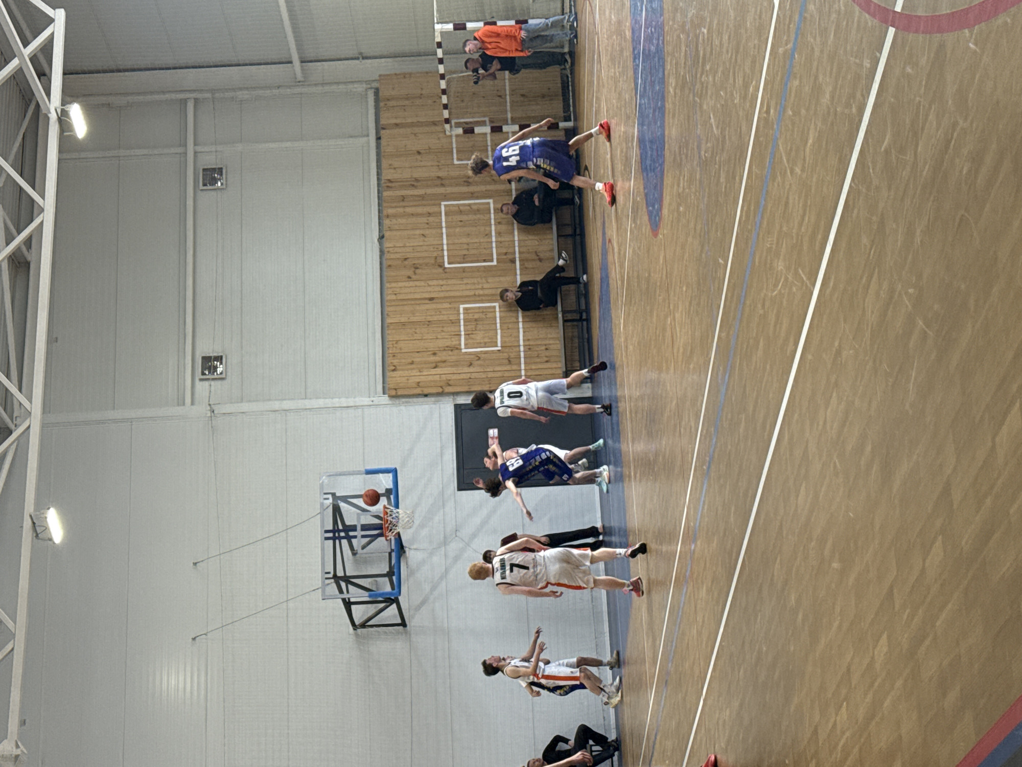 The image shows a basketball game in progress in a gymnasium. The gymnasium has a light-colored wall with a basketball hoop and a visible basketball going through the hoop. Several players wearing different colored jerseys are scattered across the court. Some spectators are visible along the sidelines. The gymnasium has white lines on the wooden court surface. The exact location in the city of Votkinsk, Russia, is not discernible from the image.