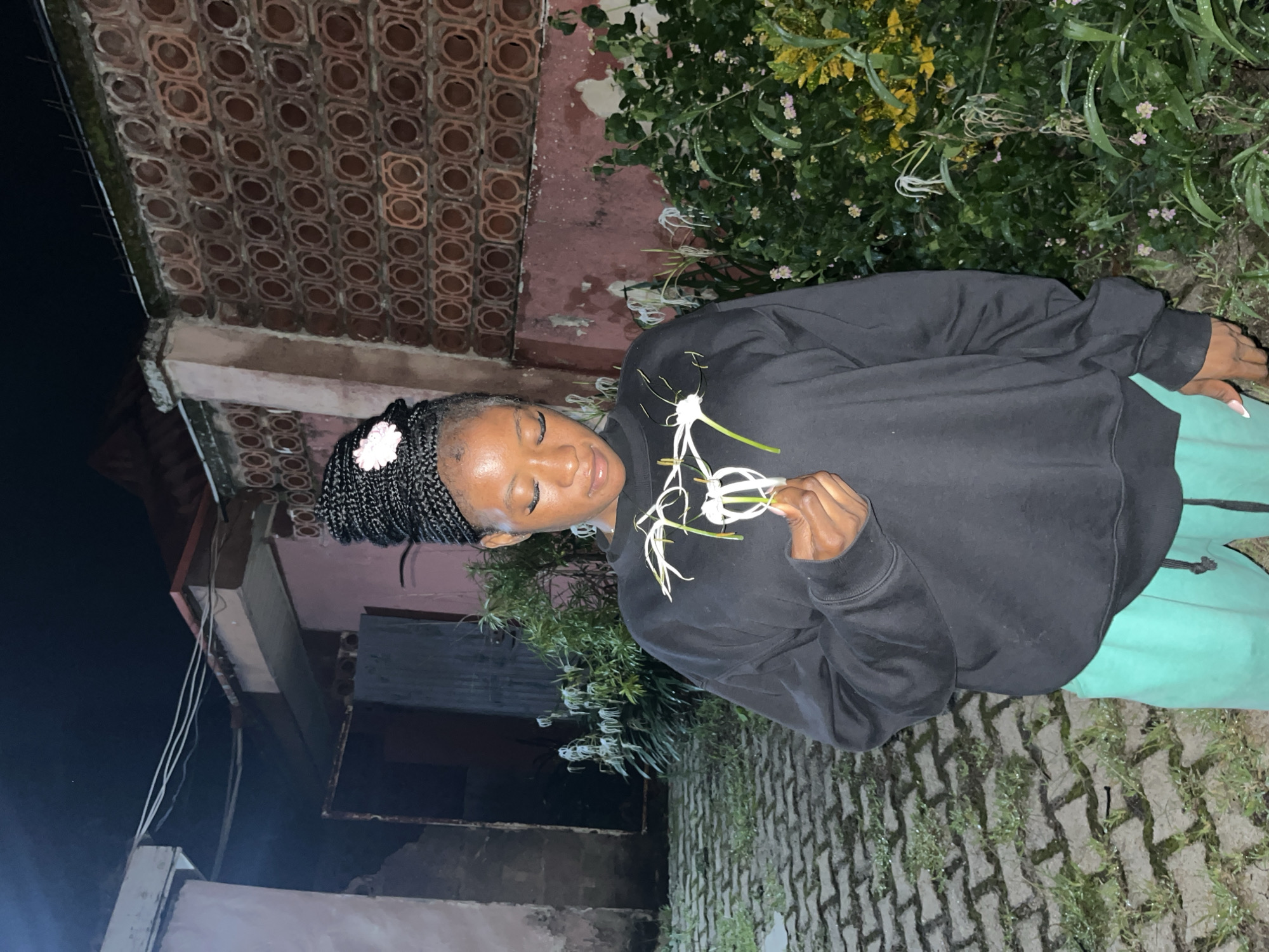 A young woman with dark skin and braided hair is standing outside at night, holding a white flower. She is wearing a black sweatshirt and teal sweatpants. The background shows a brick wall with a circular pattern and some greenery. The ground is paved with interlocking bricks and patches of grass. Overhead, there are wires and a dark sky. The overall scene suggests an outdoor setting in a residential area. The location is identified as Jos, Nigeria.