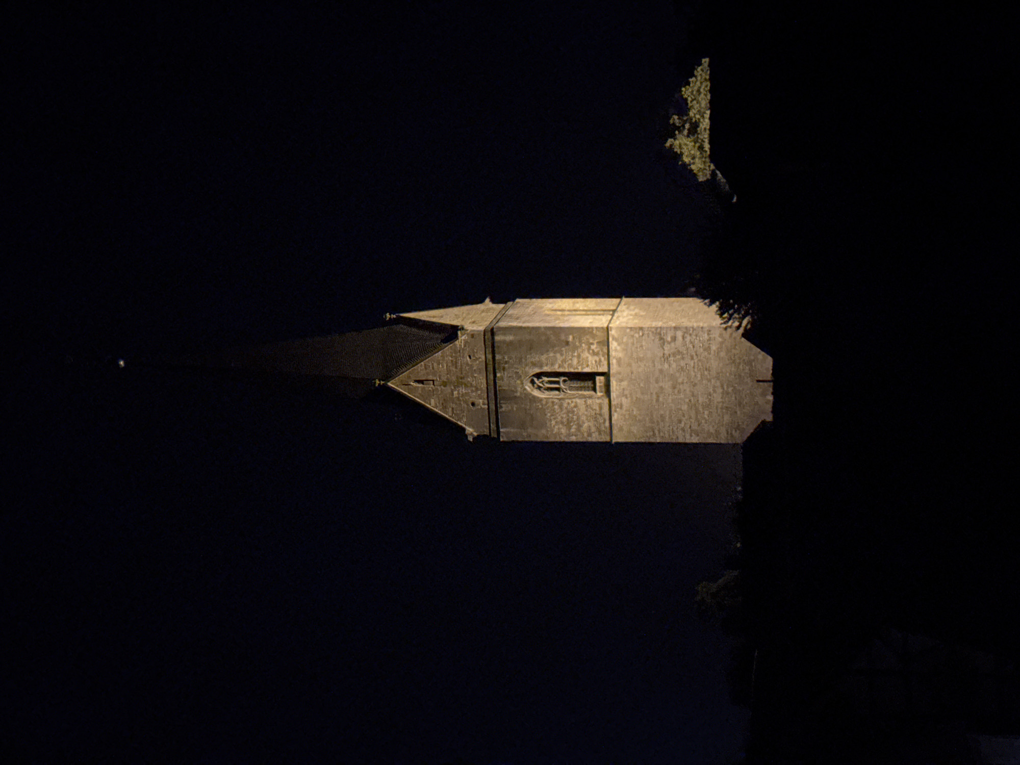 The image displays a nighttime reflection of a building, likely a church steeple, in dark water. The building is constructed of stone and features an arched window with intricate detailing. The steeple tapers to a point, and its silhouette is illuminated by artificial light, creating a contrast against the deep blackness of the night sky. The reflection in the water is distorted but clearly visible, suggesting calm or gently rippling water. To the right of the building, a tree with dense foliage is also partially visible, its branches illuminated from below and casting shadows. The overall mood is serene and slightly mysterious, enhanced by the low-light conditions. No people are visible in the frame. The time of day is definitively night, and the lack of any other environmental cues makes it impossible to determine the exact location or weather conditions, other than the presence of still water.