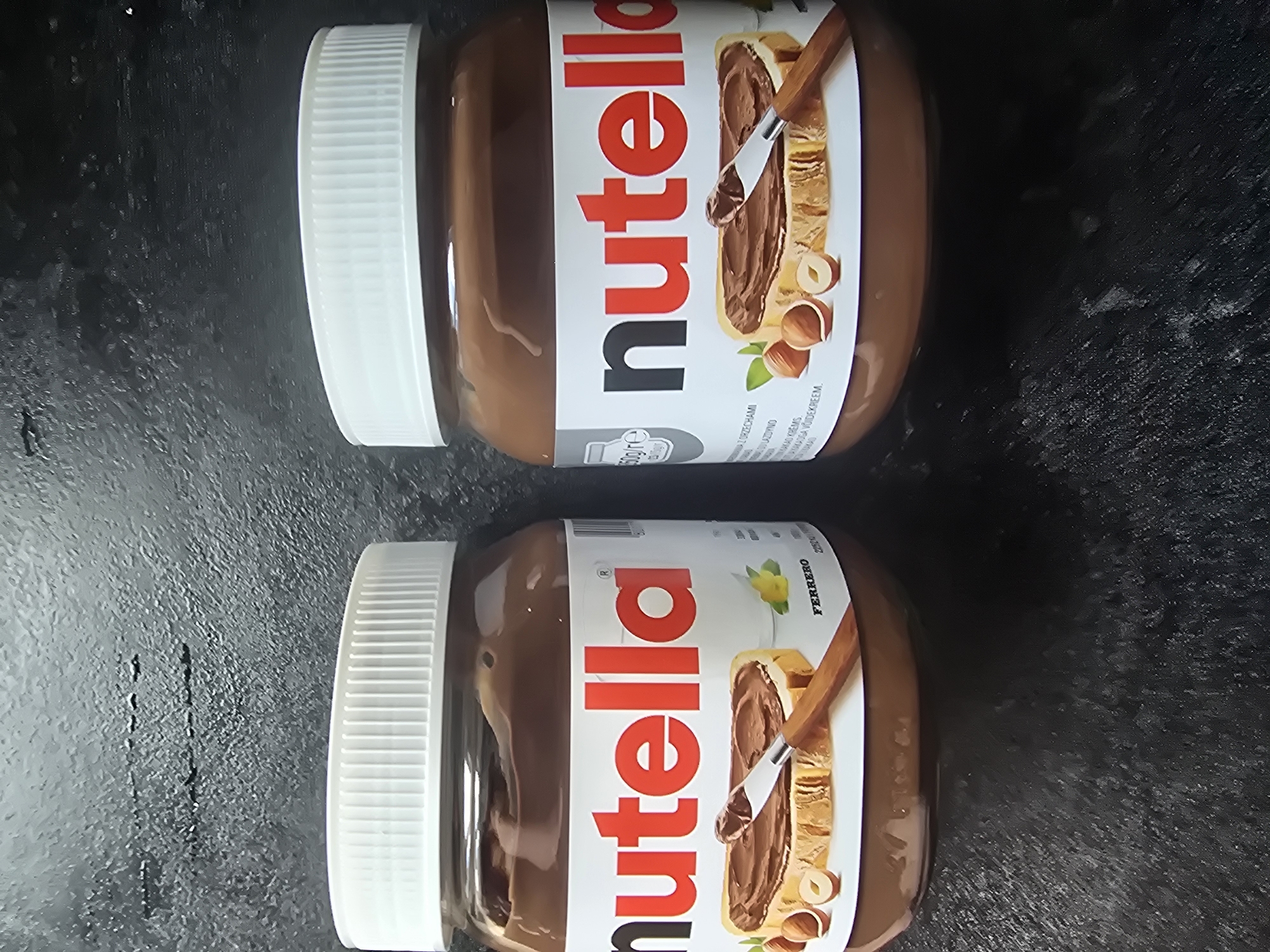 Two jars of Nutella are placed side-by-side on a dark, textured surface. Both jars have white lids and labels featuring the "nutella" logo in red and black, with an image of a slice of bread spread with Nutella, hazelnuts, and a knife. The jar on the left is slightly more in focus and shows the "FERRERO" brand name. The jar on the right has a "650g/e" label visible. The background is dark and out of focus, suggesting an indoor setting.