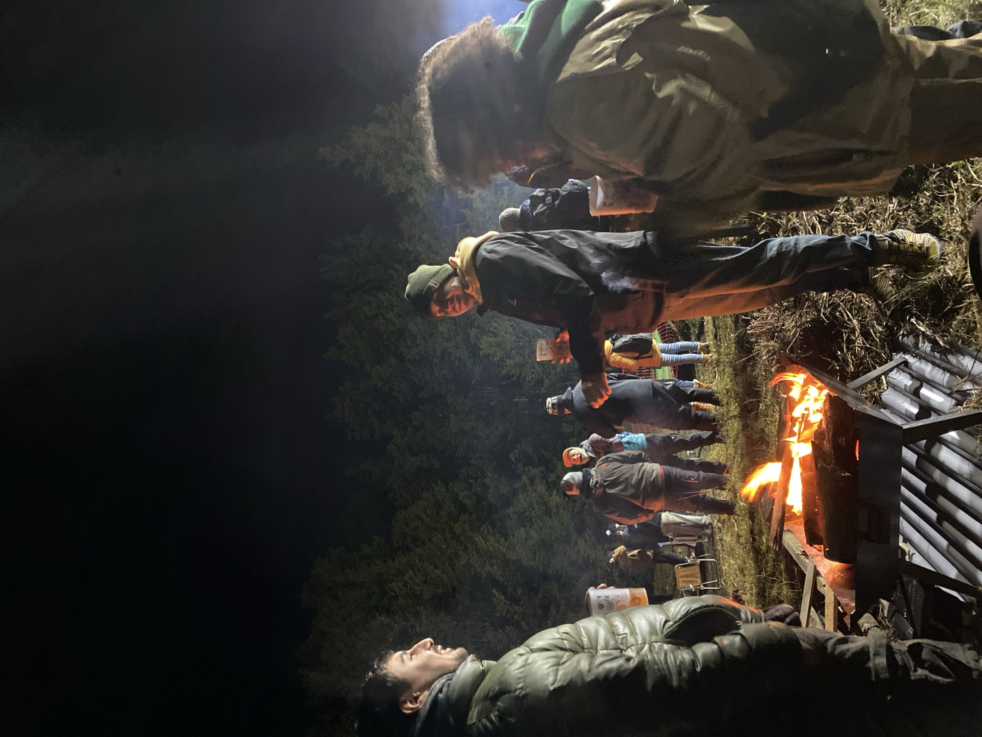 At night, an outdoor gathering surrounds a burning fire pit constructed of metal grates. The group consists of approximately ten individuals, their faces illuminated by the fire's glow. All are dressed in warm winter clothing, with hats and jackets. In the foreground, a person in a green puffy jacket reclines and looks upwards. Behind them and around the fire, other individuals stand holding cups and talking. Trees are visible in the background, shrouded in darkness. The location is likely rural, outside of the city, as evidenced by the darkness and vegetation. There is no indication in the photograph to confirm the location is Ushuaia, Argentina.