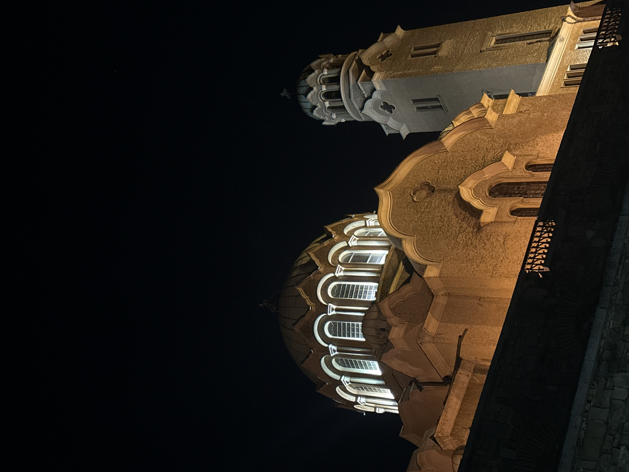 At night, the Cathedral of the Assumption in Veliko Tarnovo, Bulgaria, is seen from a low angle. The dome of the cathedral is lit, and its windows are visible. A tower-like structure is above and behind the dome. The sky is dark. A portion of an adjacent building is visible on the right.