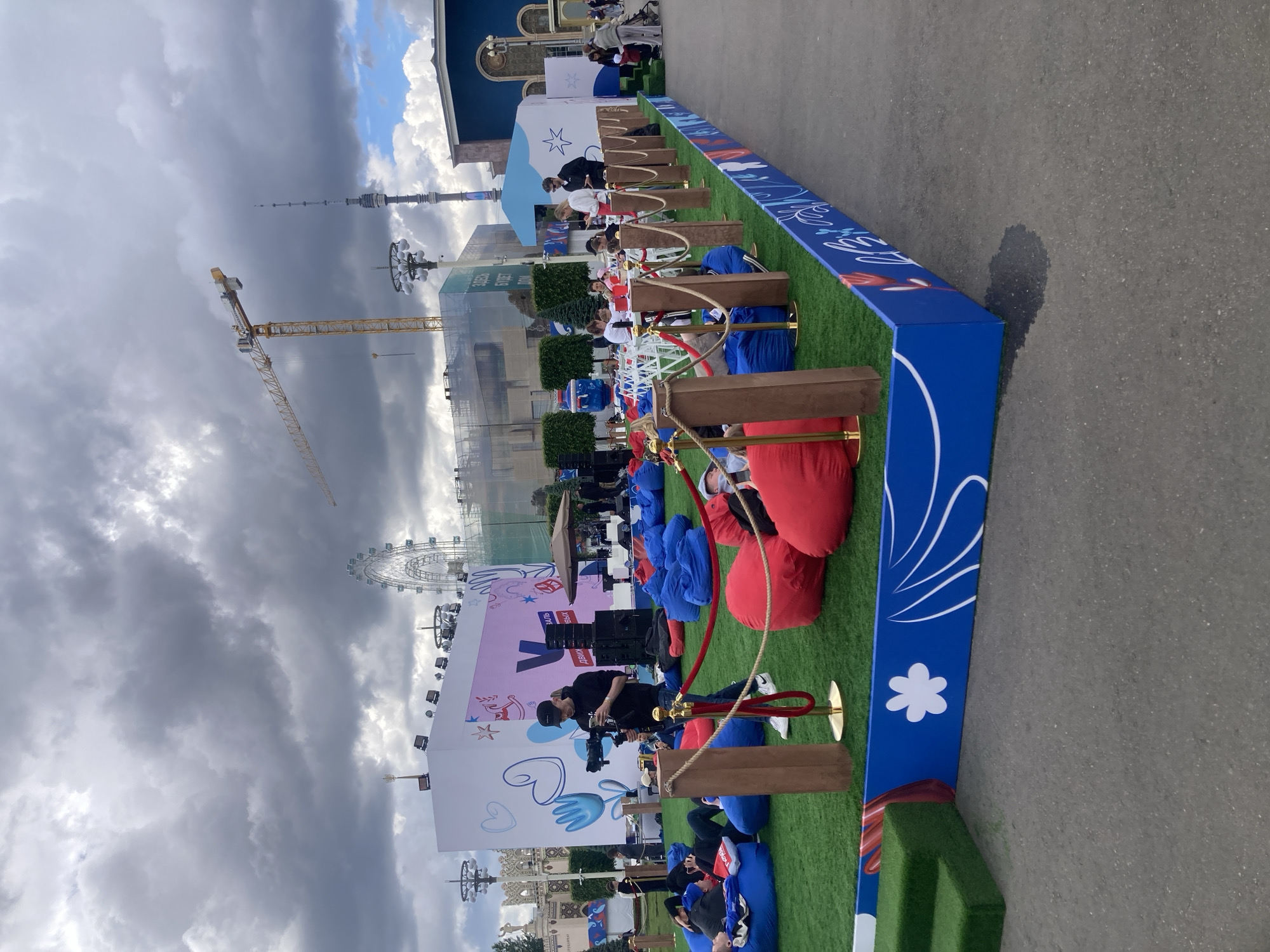 The image shows an outdoor event setup in Moscow, Russia, discernible by the presence of the Ostankino Tower and a Ferris wheel in the background. In the foreground, there's a designated area with artificial turf, bean bag chairs in blue and red, and wooden dividers. The event space is bordered by a blue and white decorative barrier. People are seated and appear to be relaxing or observing something. A camera operator is capturing footage. Construction is visible in the background, indicated by a crane. The sky is cloudy and overcast.