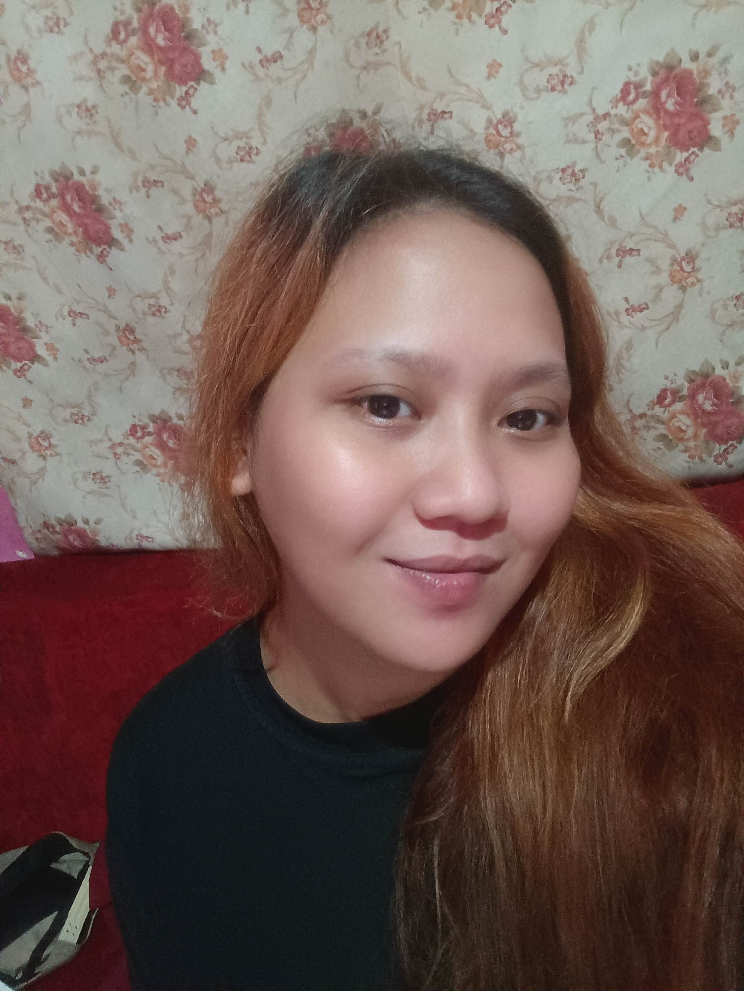 The image shows a close-up, head-and-shoulders selfie of a young woman with light brown hair, wearing a black t-shirt. She is positioned in front of a floral wallpaper, with a red sofa visible behind her. A bag is partially visible on the left side of the image. The scene appears to be indoors, likely in a residential setting within Depok, Indonesia. The lighting suggests it was taken in a room with ambient light.