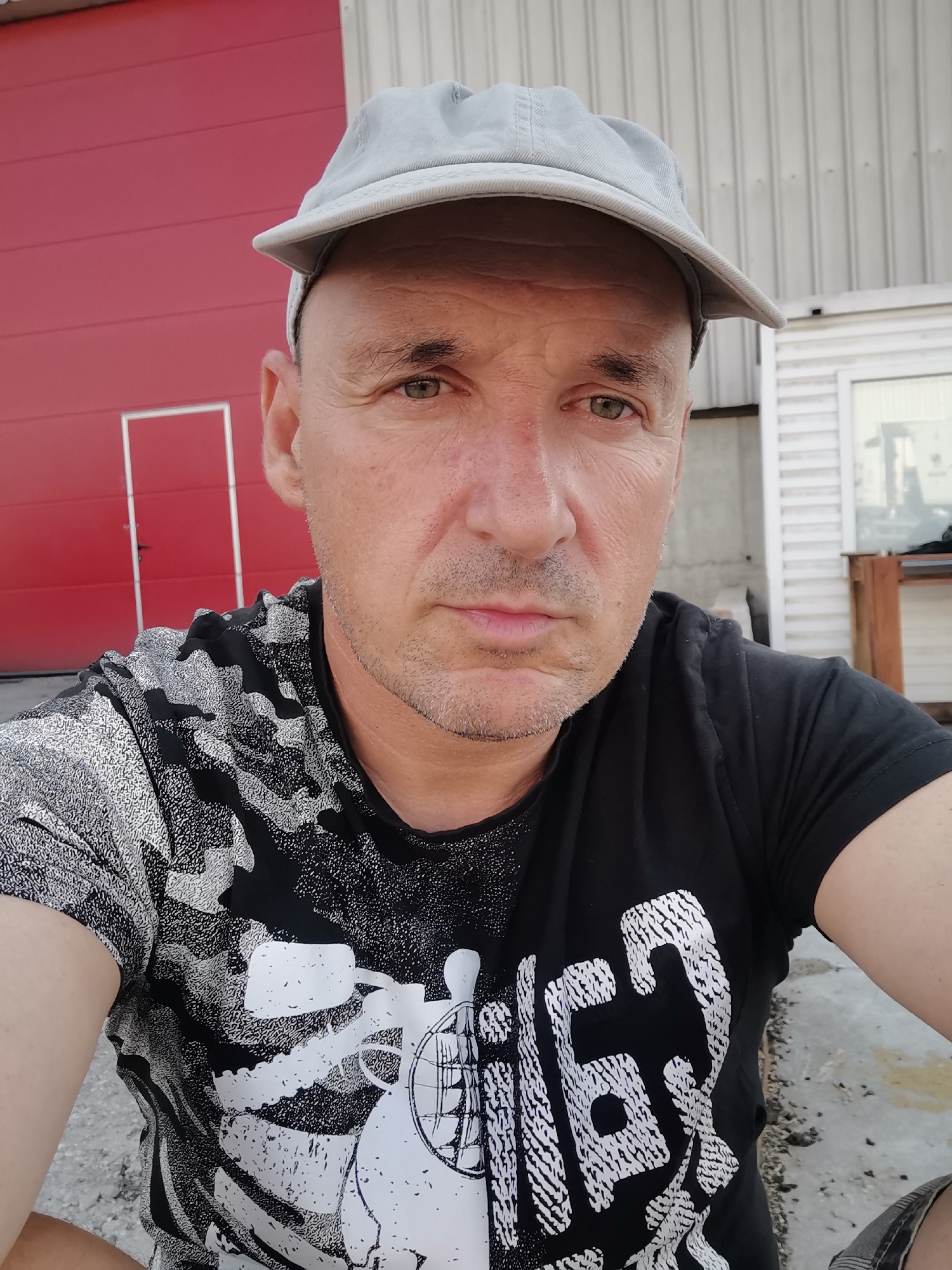 A man wearing a grey baseball cap and a black t-shirt with a white graphic design is pictured in an outdoor setting. He has short, greying hair and light-colored eyes. The background features a large red garage door with a white framed entrance on the left, and a light-colored corrugated metal building with a window on the right. The ground appears to be a rough, gravelly surface. The image is a selfie, taken from a medium close-up perspective.