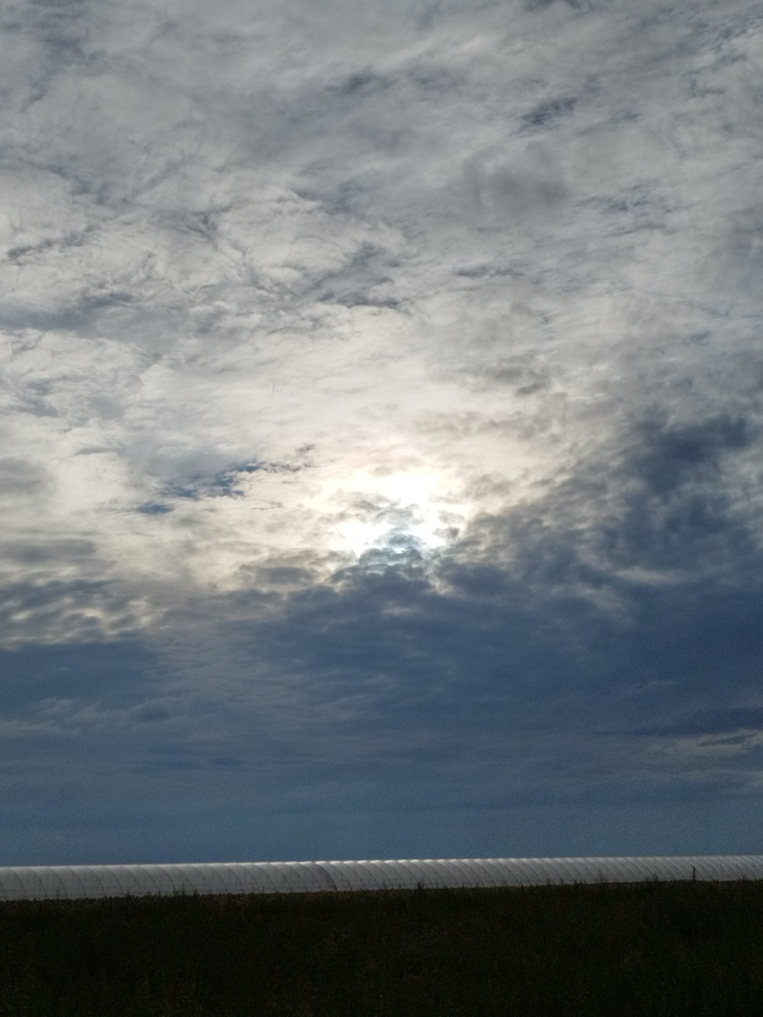 The image depicts a wide, horizontally oriented scene under a cloudy sky. The sky occupies the upper two-thirds of the frame and features a mix of dark and light gray clouds. The sun is partially obscured by clouds in the upper center, creating a bright, hazy glow. Below the sky, a dark, horizontal band of ground is visible, topped by a row of translucent, arched structures that appear to be greenhouses. The ground in front of the structures is dark and appears to be covered in vegetation or soil. The scene suggests an outdoor agricultural setting, possibly in Băleni-Sârbi, Romania, given the presence of greenhouses. There are no people or animals visible in the image. The lighting is diffused due to the cloud cover, with the sun's glare being the brightest point.