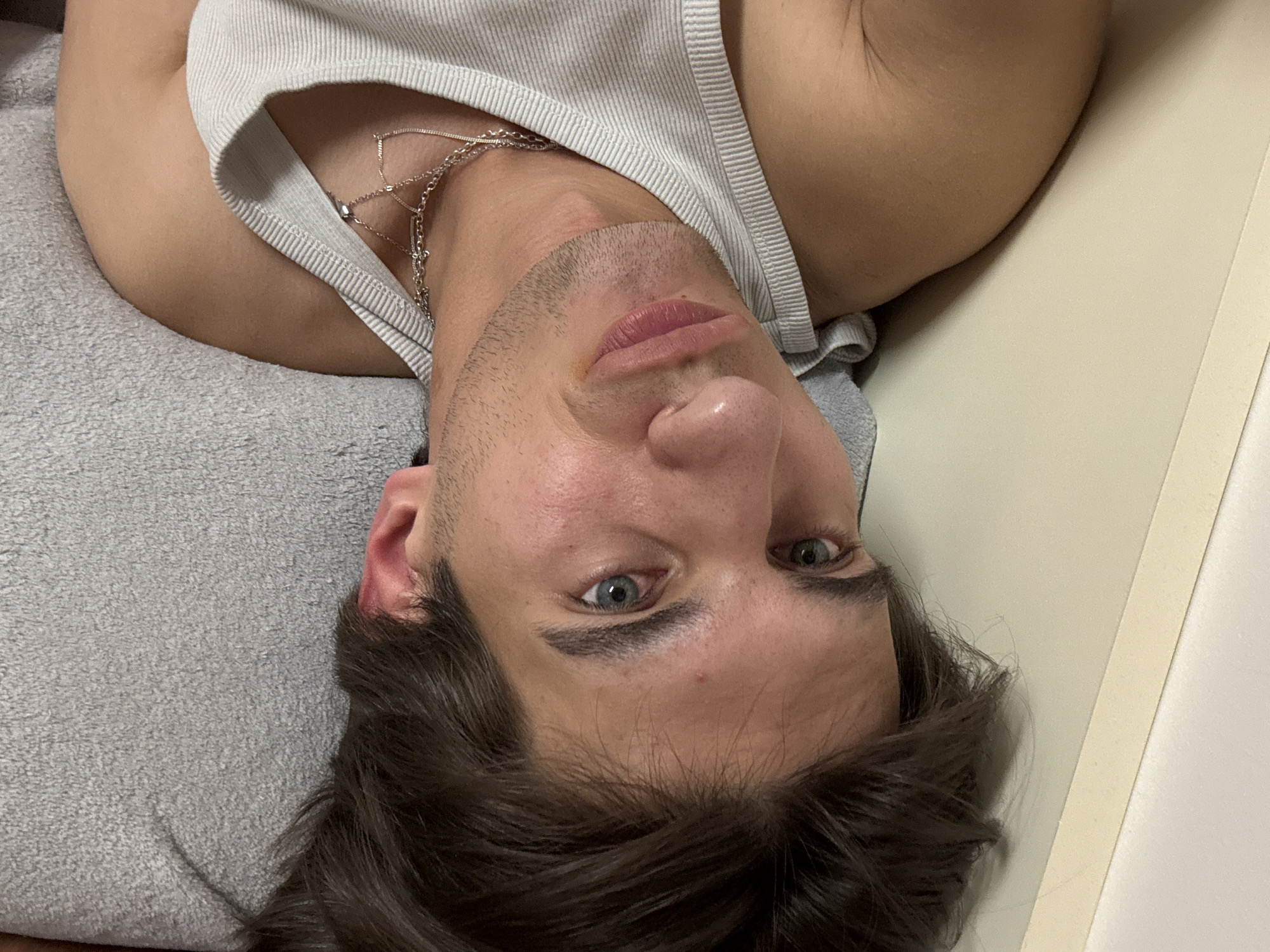 A man is lying down, possibly on a bed or couch, with his head resting on a gray towel-like surface. He is wearing a white tank top and a silver necklace. His hair is dark brown and his eyes are blue. The background includes a light-colored wall or surface. It is not possible to determine the location as Sofia, Bulgaria from this image alone.