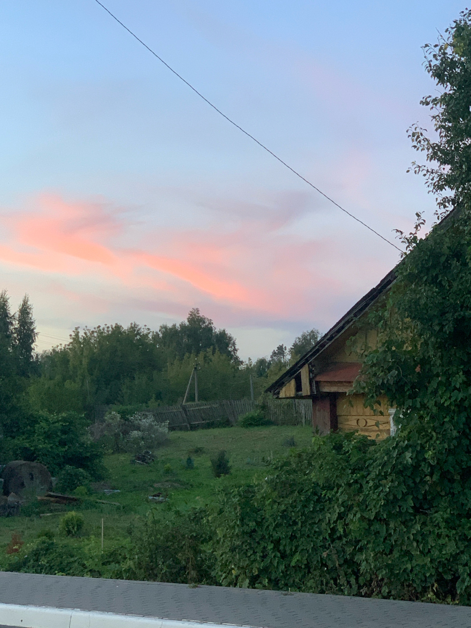 This is a medium-shot, eye-level, horizontal photograph taken outdoors in Kapyĺ, Belarus, during twilight. The image depicts a rural scene featuring a yellow wooden house partially obscured by dense green foliage in the foreground and midground on the right. The house has a dark red door and a sloping roof. Behind the house, a weathered wooden fence runs diagonally across the midground. Beyond the fence is a landscape of rolling green hills dotted with trees. The sky above is a pale blue, transitioning to a soft pink hue due to sunset clouds. A thin, dark power line stretches diagonally across the sky from the top left corner towards the mid-right. In the far left midground, a large, round, stone object is visible amongst some debris. The overall atmosphere is peaceful and serene.