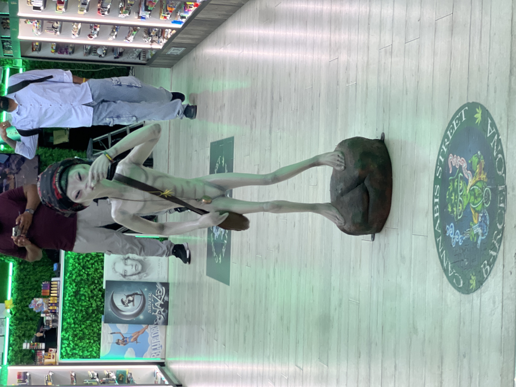 The image depicts the interior of a retail establishment, possibly a cannabis dispensary, located in Barcelona, Spain. A life-sized, pale green statue of an alien is positioned in the foreground, standing on a dark, rocky base. The alien statue is unclothed and has large eyes and a slender build. It is wearing a dark head covering and a dark satchel slung across its chest.
In the background, two individuals are visible. On the left, a person wearing a maroon t-shirt and light gray cargo pants is looking at their phone. On the right, another person is wearing a light blue collared shirt, light gray cargo pants, and a black backpack, and appears to be walking away from the camera.
The environment features a polished wooden floor. Several posters are displayed on the wall behind the alien statue, featuring imagery related to cannabis products, including a depiction of a woman on a swing labeled "BLUEDREAM," a crescent moon with a figure labeled "SILVER HAZE," and a portrait of a woman labeled "WHITE WIDOW." Shelves stocked with various products are visible along the right wall of the establishment. A circular logo on the floor in the foreground displays the text "CANNABIS STREET BARCELONA." The overall lighting is a soft, greenish hue.