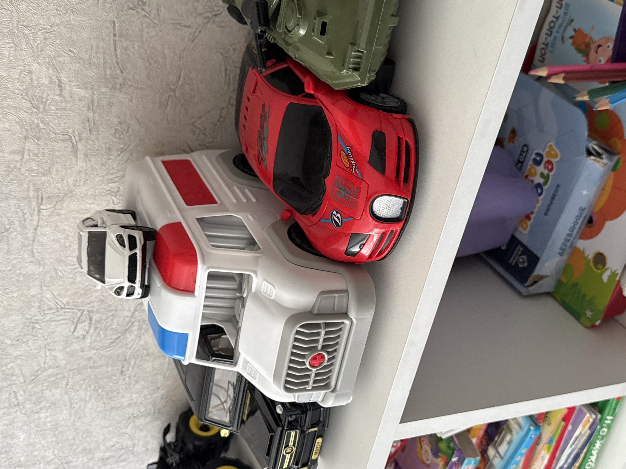 A close-up, eye-level shot shows a collection of toy vehicles and books on a white shelf. In the foreground, a bright red toy sports car is positioned next to a green toy tank. Behind the sports car is a white toy ambulance with red and blue accents on its roof. To the left of the ambulance is a black toy SUV. A small white toy car is perched on top of the ambulance. The background features a textured, light-colored wall. Below the shelf, partially visible, are stacks of colorful children's books and a purple object. The toys appear to be in good condition, and the overall scene suggests a child's play area. The location is noted as Яромінскі сельскі Савет, Belarus, which is a rural administrative division.