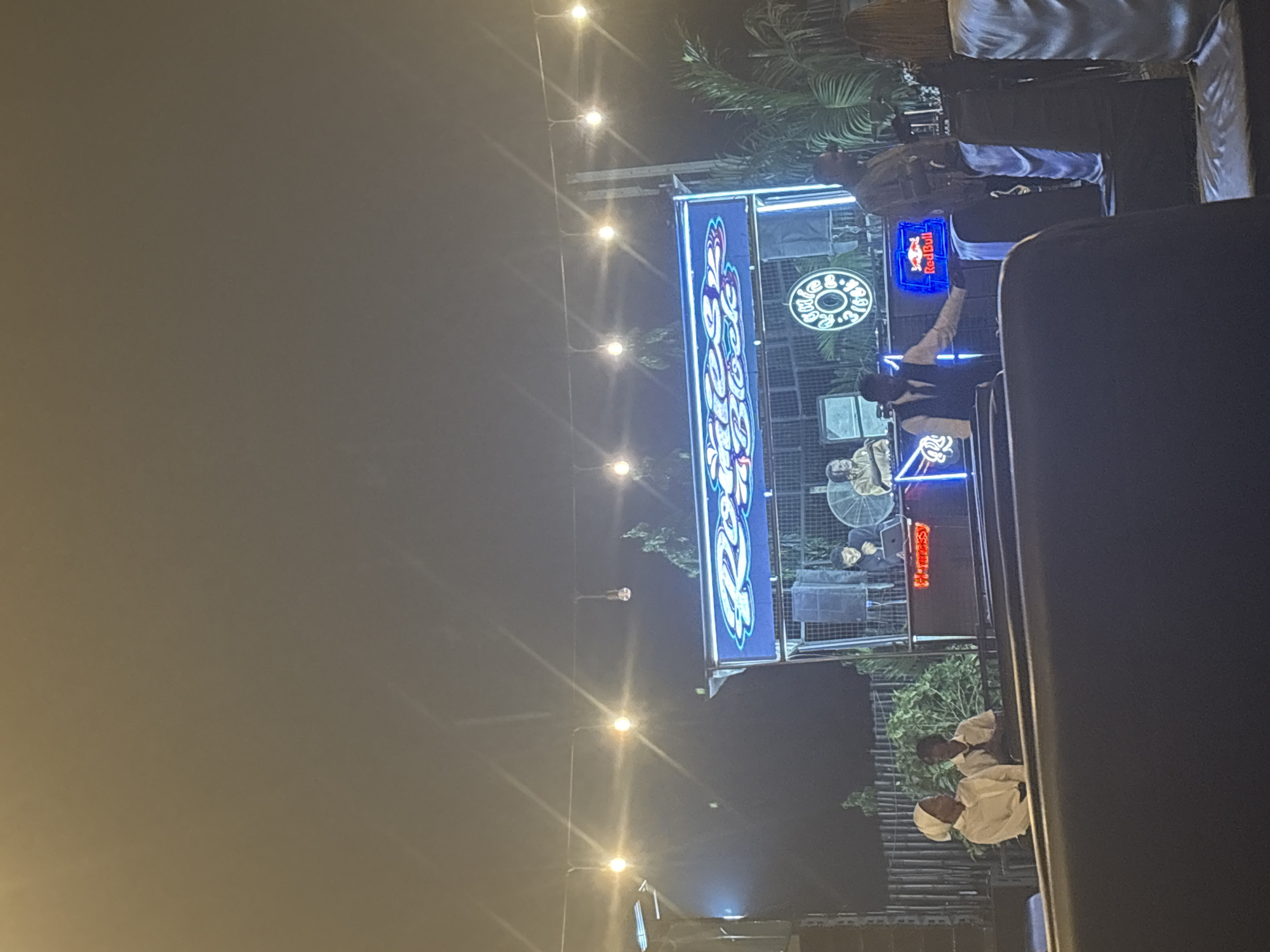 The image shows an outdoor restaurant or event space at night, likely in Abuja, Nigeria, indicated by the "Lee's Bistro" sign. The area is illuminated by overhead string lights. A large, brightly lit sign for "Lee's Bistro" is visible, alongside a Red Bull sign. People are present; some appear to be staff in white uniforms, while others are customers. The environment includes greenery, suggesting an outdoor setting. Some people are standing around a DJ booth, suggesting the presence of live music or entertainment. There is also a table with people sitting down and eating.