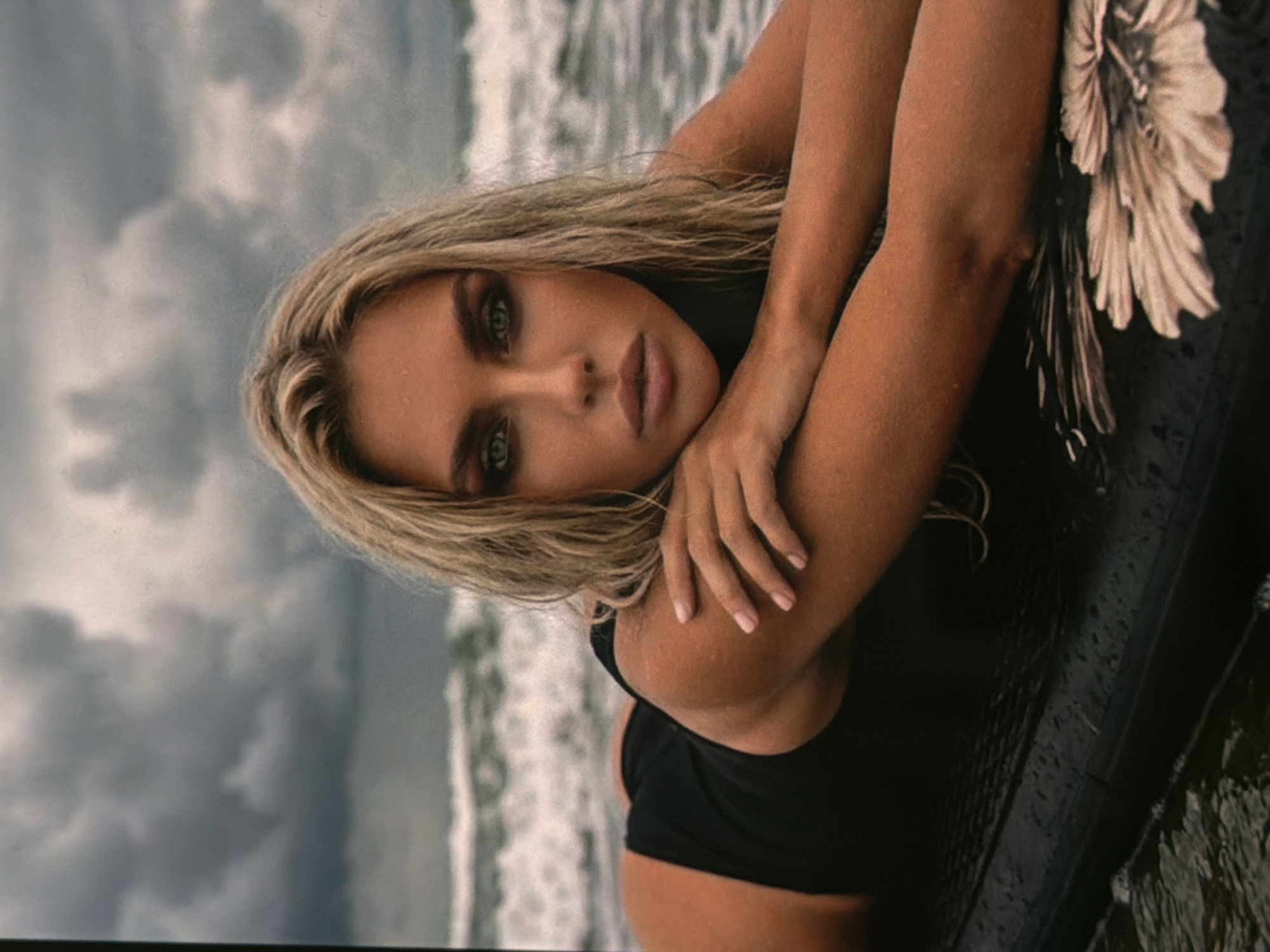 An athletic woman with blonde hair and green eyes, wearing a black swimsuit, lies on a surfboard in the ocean. The waves are choppy, and the sky is overcast with dark clouds. The surfboard has a floral pattern on its tail. The scene appears to be a candid shot, possibly taken during a photoshoot.