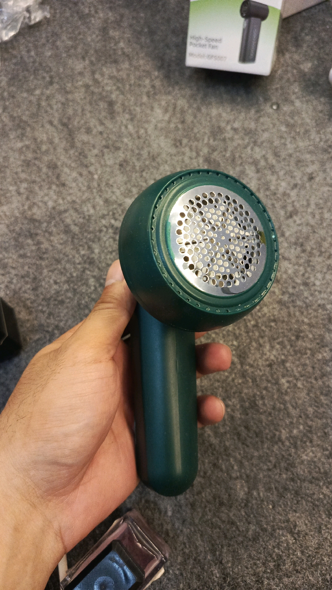 A hand holds a teal fabric shaver with a circular metal grate containing many small holes. The background is a grey, textured surface. In the upper right corner, a portion of a white box is visible with text that reads "High-Speed Pocket Fan" and "Model: OP0207". A small, clear rectangular object with a blue interior is in the lower left of the frame. The scene is indoors, and the overall impression is of a product review or unboxing context. There is no indication of a specific location in Babadan, Indonesia.