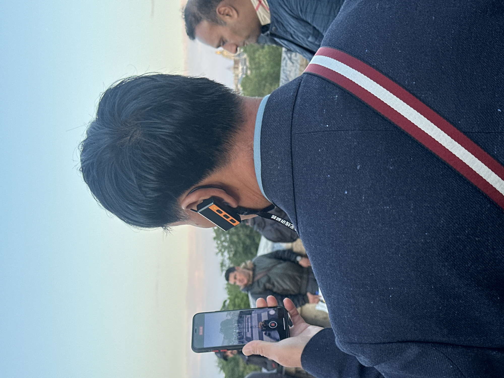 An individual with dark hair is holding a smartphone in landscape orientation, taking a video or photograph of a group of people outdoors. The person wears a dark blue jacket with a red and white stripe along the shoulder strap. An earpiece is worn in the ear. Other people can be seen in the background, with a hint of Parisian architecture in the upper part of the frame. Given the architecture, the location is likely Paris, France.