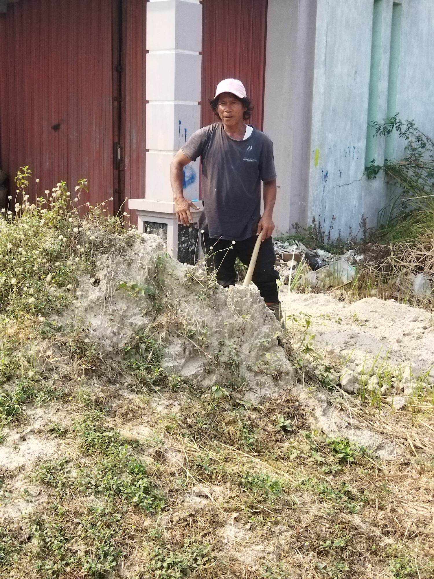 A man wearing a pink baseball cap and a dark t-shirt stands outdoors in what appears to be a construction or landscaping area in Suka Sari, Indonesia. He is holding a shovel in his right hand, with the blade resting on a pile of dirt and vegetation in front of him. The man is looking towards the camera with a neutral expression. Behind him, to his left, is a building with a large, reddish-brown corrugated metal door. To his right is a light-colored wall with vertical panels and some graffiti. The ground is covered in dry grass, dirt, and some scattered debris. A small mound of dirt and plants is positioned prominently in the foreground, with the man standing behind it. The overall scene suggests an outdoor work environment with elements of both construction and overgrown vegetation.