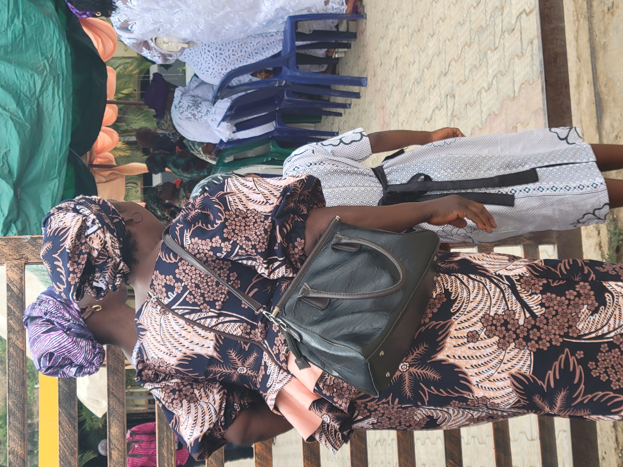 A woman sits outdoors in Suleja, Nigeria. She wears a patterned dress and headwrap, and a black purse hangs over her shoulder. Another person stands behind her, wearing a white dress with a black belt. Stacks of blue plastic chairs and people under a green tarp are visible in the background. The ground is paved. The scene appears to be at an event or gathering.